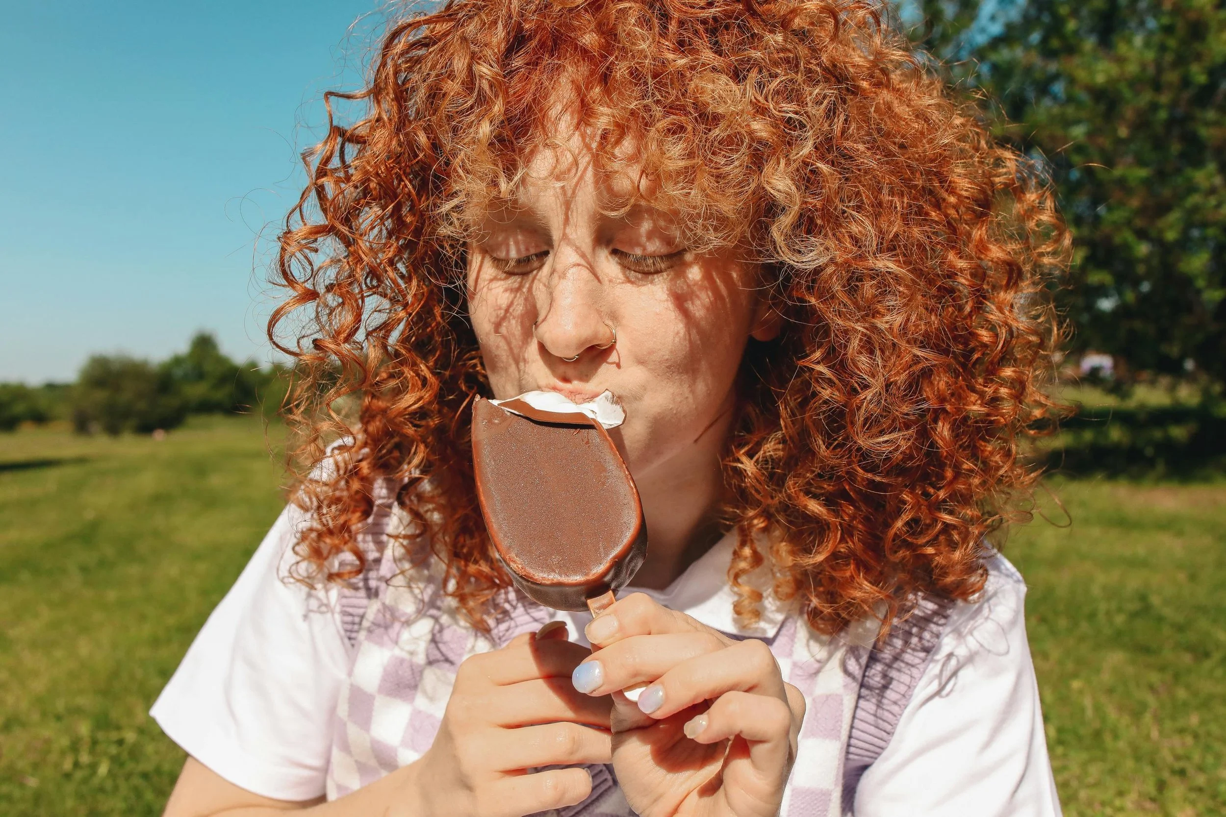 Woman with red, curly hair enjoying a chocolate covered yogurt bar outside in a field of green grass.