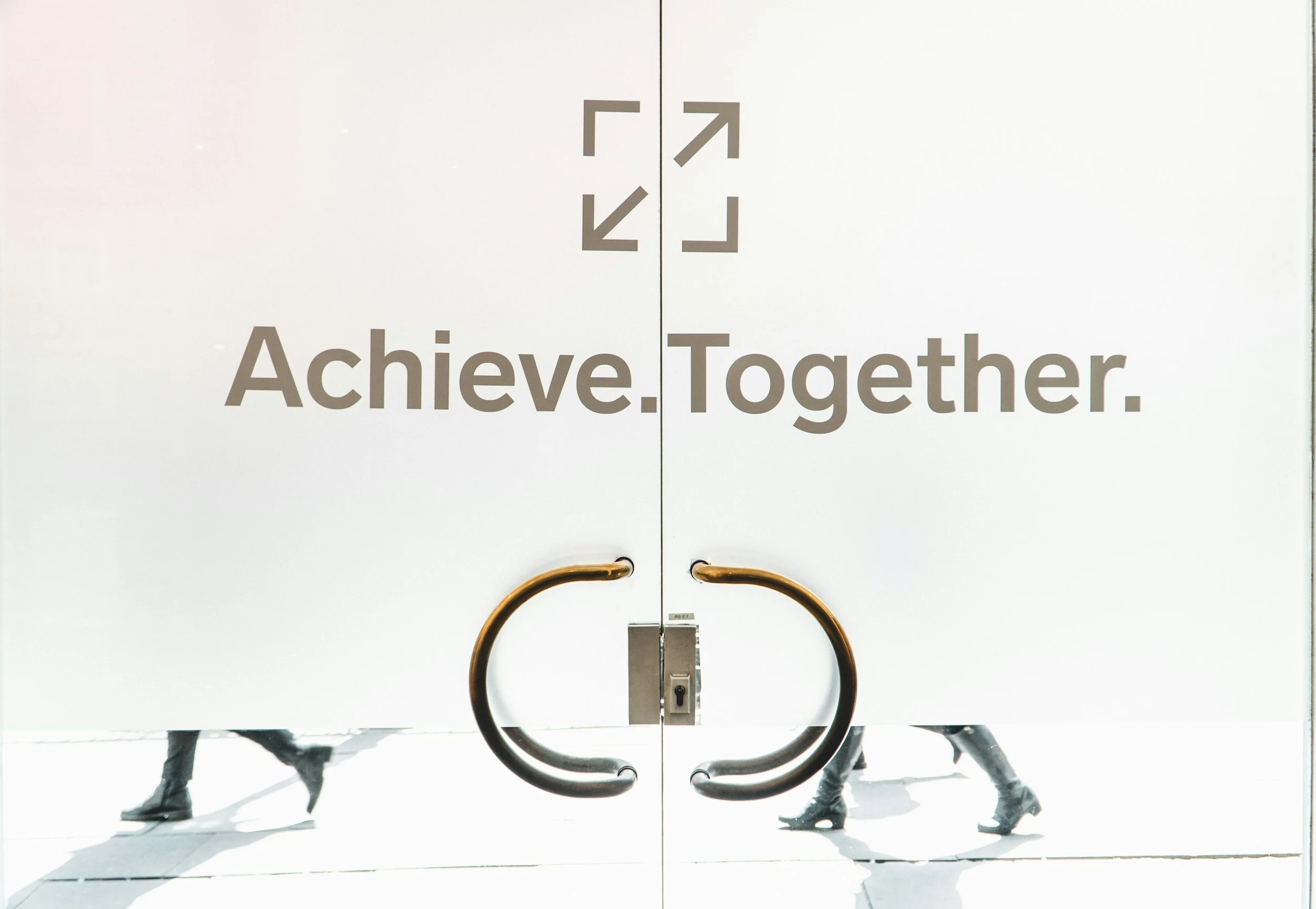 White double doors with the words 'Achieve. Together.' printed on them and two large, curved door handles. Reflection of pedestrians walking past on the sidewalk behind the doors.