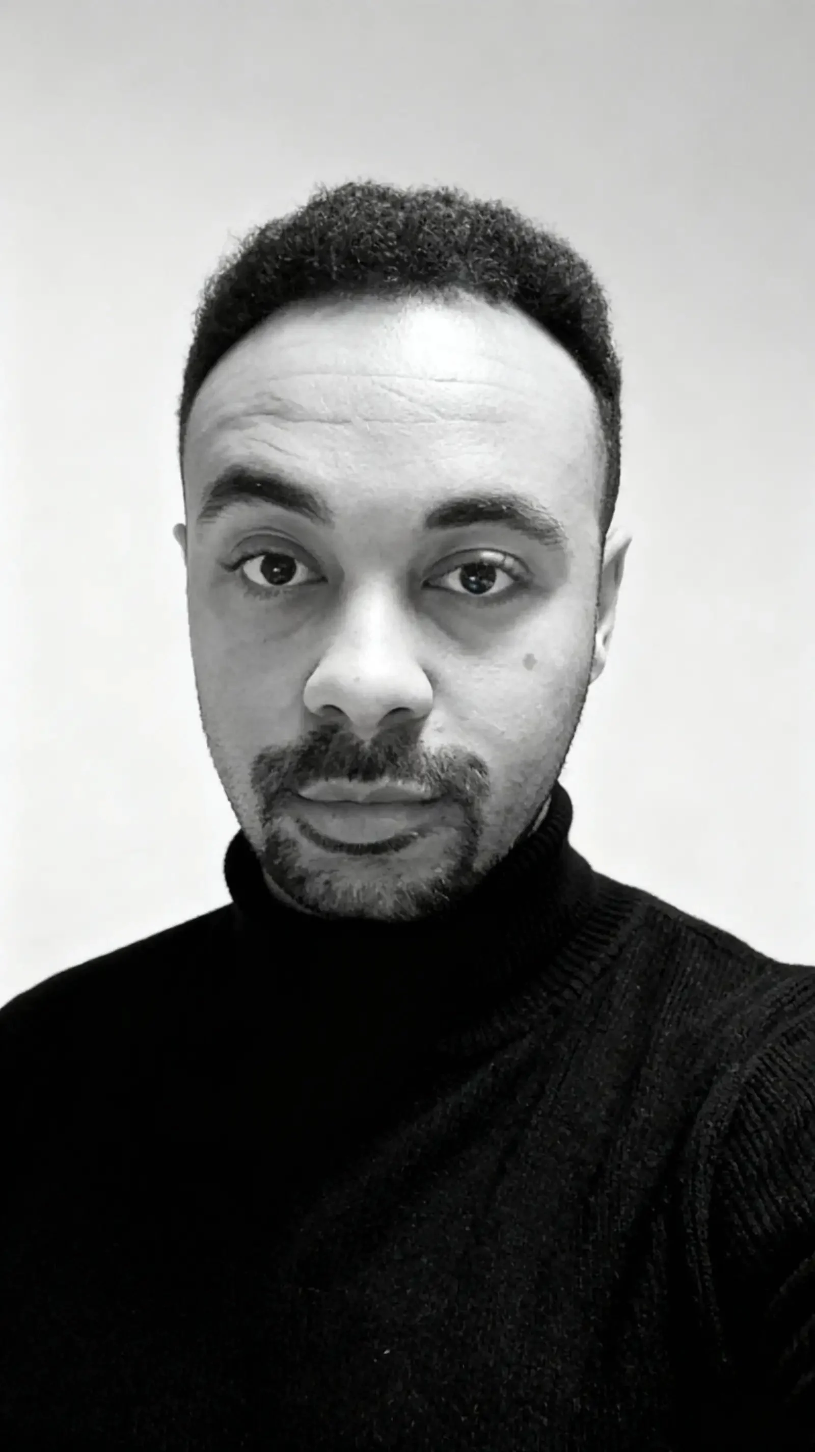 Black and white close-up photo of a man with short curly hair, beard, and mustache, wearing a dark turtleneck sweater, looking directly into the camera.