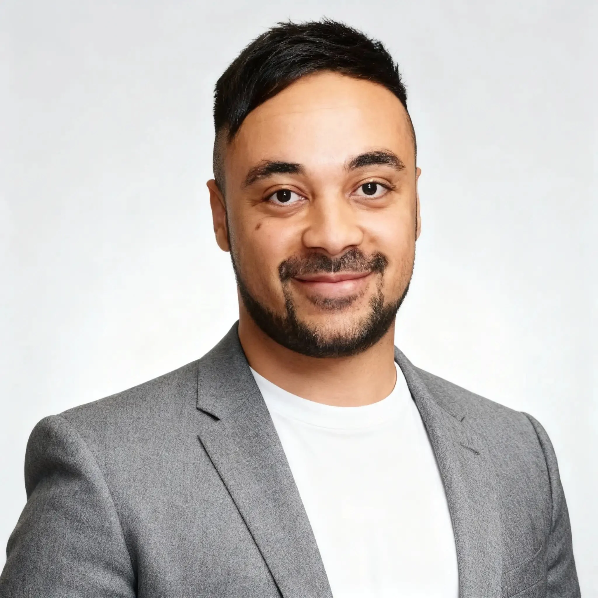 A man with short black hair and a beard wearing a gray blazer and white t-shirt, smiling against a plain white background.