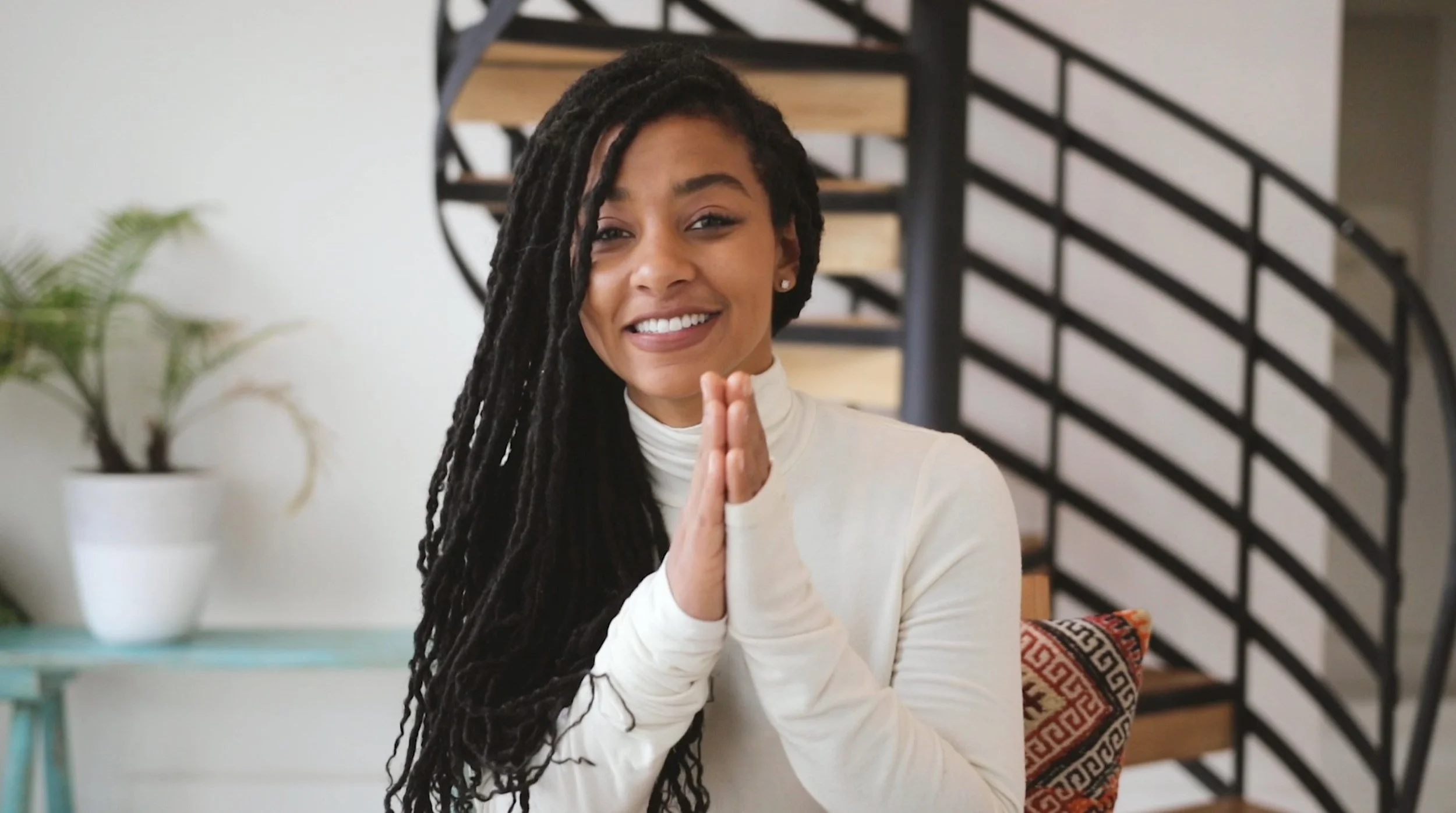 A woman with long braided hair smiles while sitting in front of a spiral staircase and a potted plant. She is wearing a white turtleneck and has her hands together, appearing friendly and relaxed.