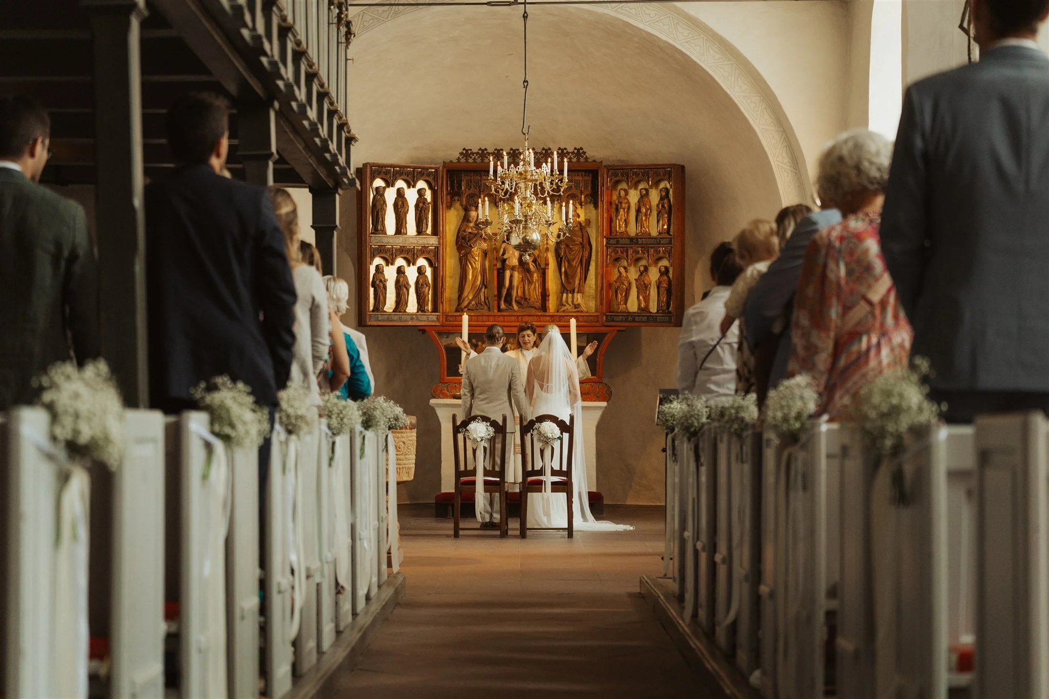 Hochzeit in der St. Severin in Keitum auf Sylt. Braut, Bräutigam und Pfarrer stehen vorne in der Mitte, umgeben von anwesenden Gästen die auch stehen, mit religiöser Ikonografie im Hintergrund und festlicher Dekoration.
