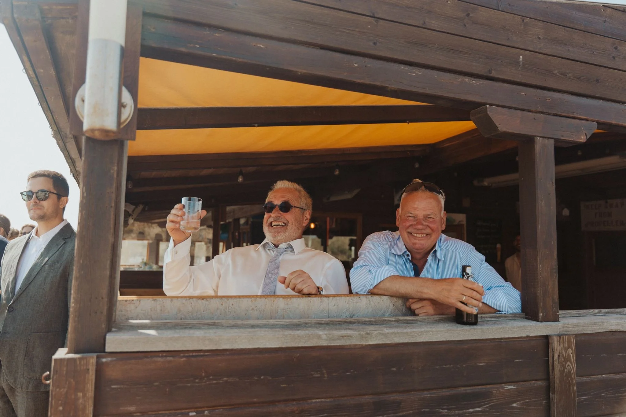 Schnappschuss von Bräutigamsvater und einem weiteren männlicher Gast bei einer Hochzeit auf Sylt, der eine hält ein Glas mit einem Getränk, während der andere lächelt. Im Hintergrund sind weitere Menschen und eine Barstruktur sichtbar.
