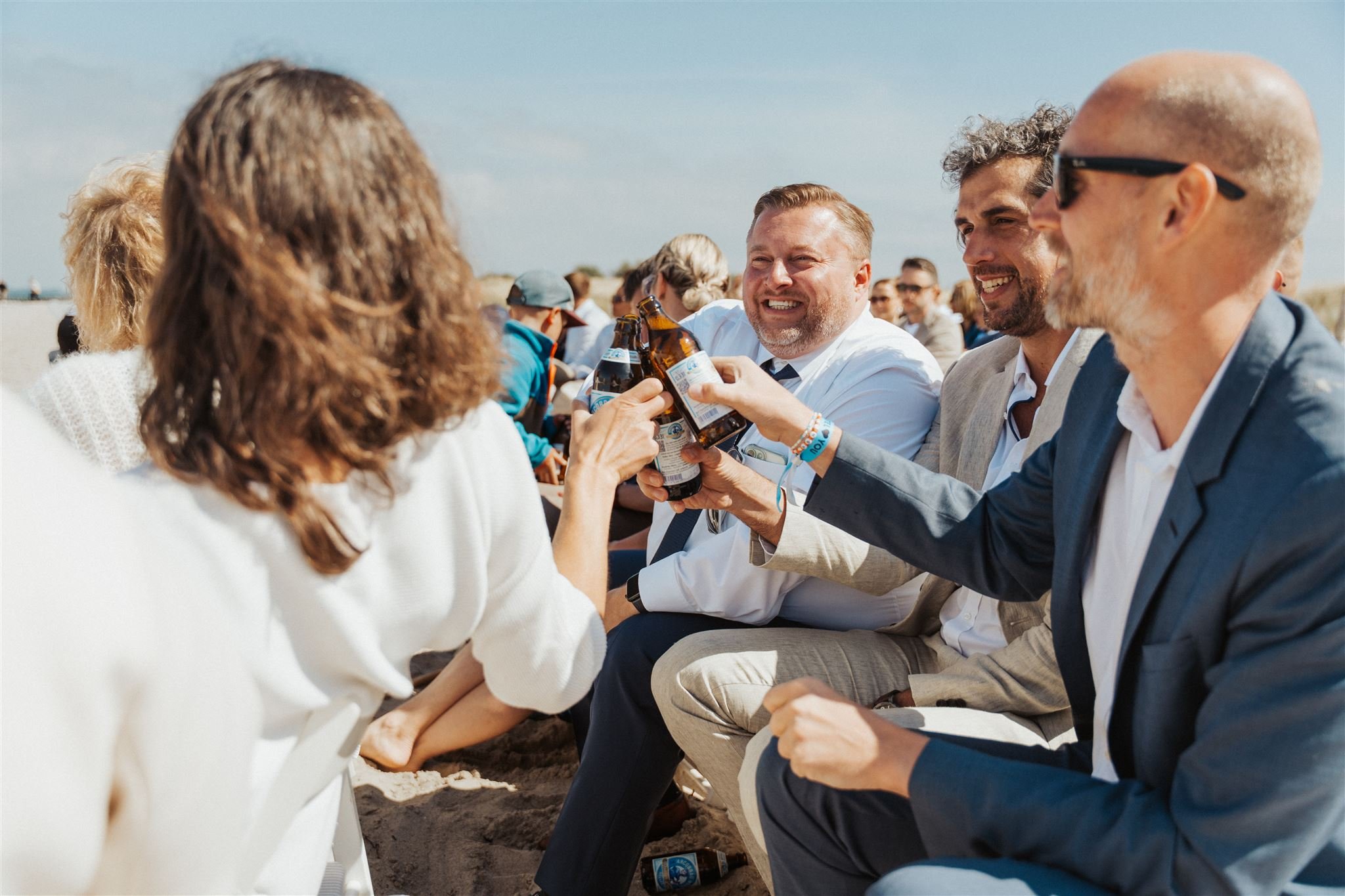 Hochzeitsgäste sitzen bei freier Trauung Strand, stoßen mit Bierflaschen an und lachen. Es ist ein sonniger Tag, sie lachen und tragen festliche Kleidung.
