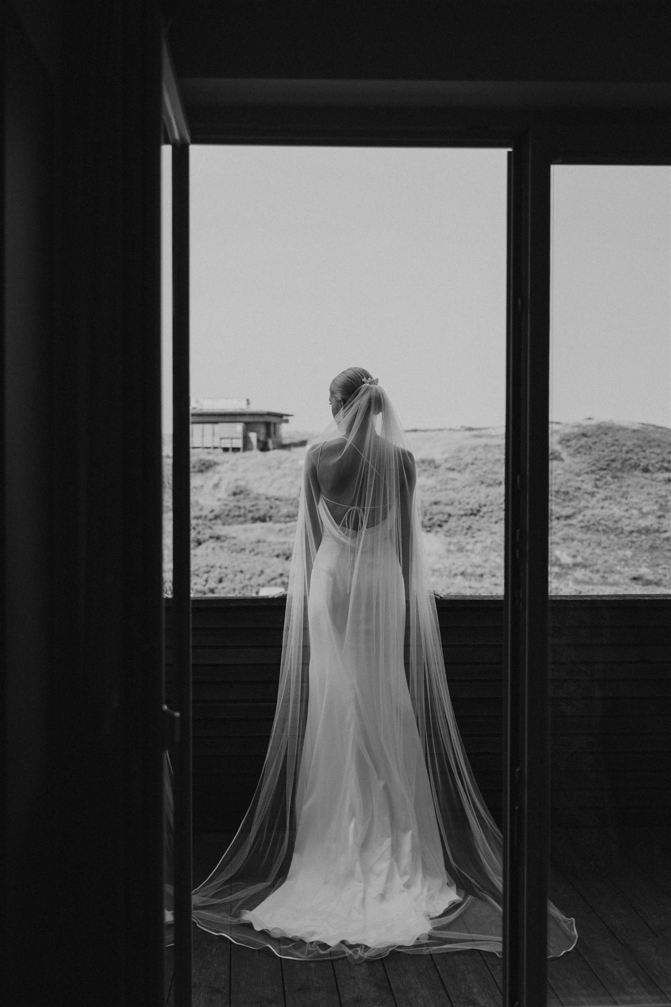 Schwarz-Weiß-Foto einer Braut beim Getting Ready, die auf einer Veranda schaut, mit Blick auf eine Landschaft. Sie trägt ein langes, fließendes Brautkleid und einen Schleier.