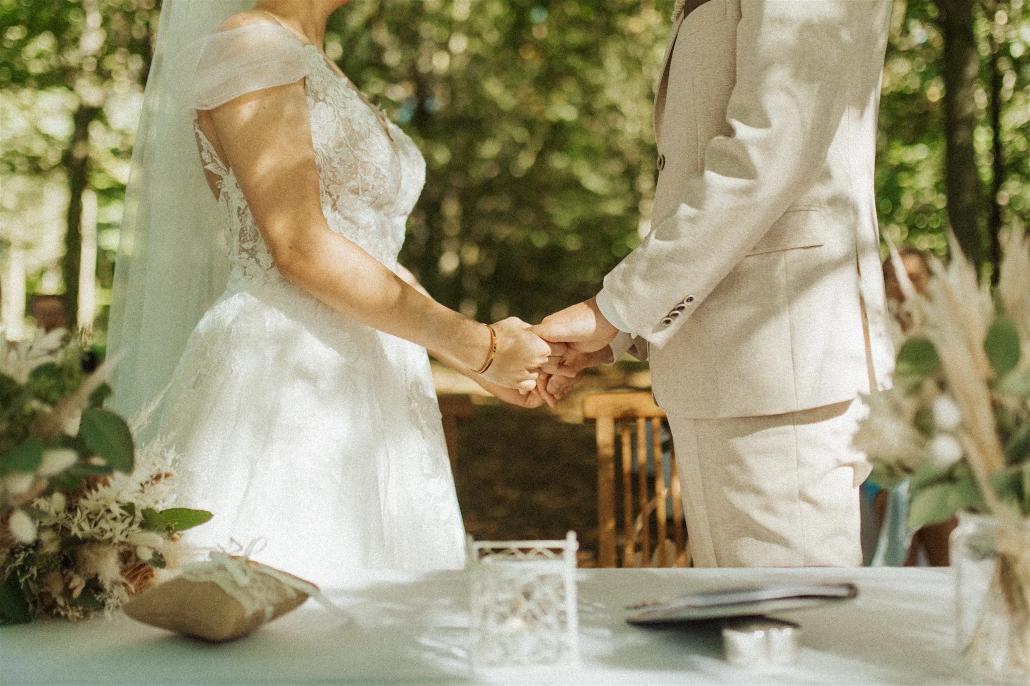 Ein Brautpaar sich hält bei einer Hochzeit im Freien, umgeben von Bäumen und Blumen, mit einem festlich gedeckten Tisch im Vordergrund.