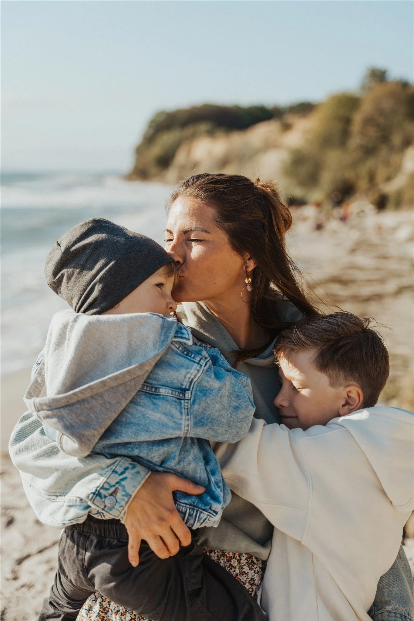 Eine Frau umarmt ihre zwei Kinder am Strand beim Familienshooting, die Kinder kuscheln sich an sie. Im Hintergrund ist die Grömitzer Steilküste zu sehen.