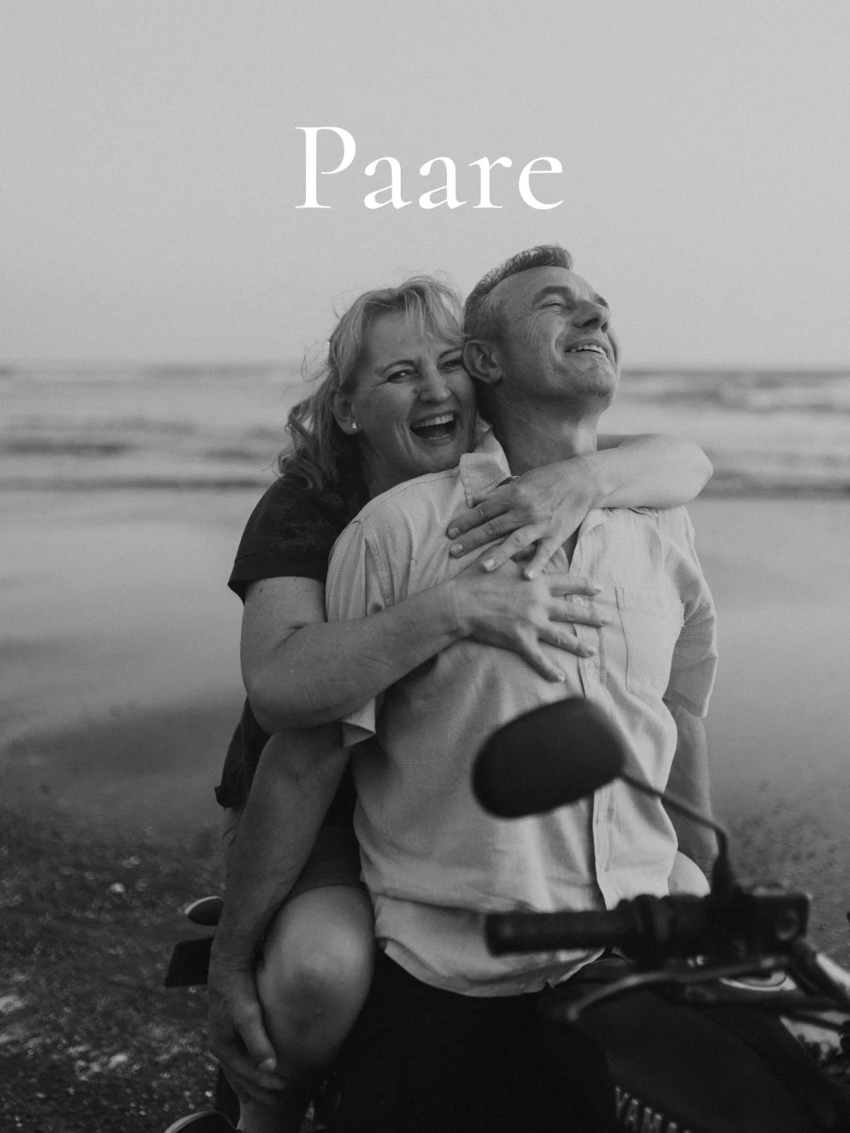 Ein lachendes Paar, das auf einem Fahrrad am Strand sitzt, die Frau umarmt den Mann von hinten, im Hintergrund das Meer, in Schwarz-Weiß aufgenommen.