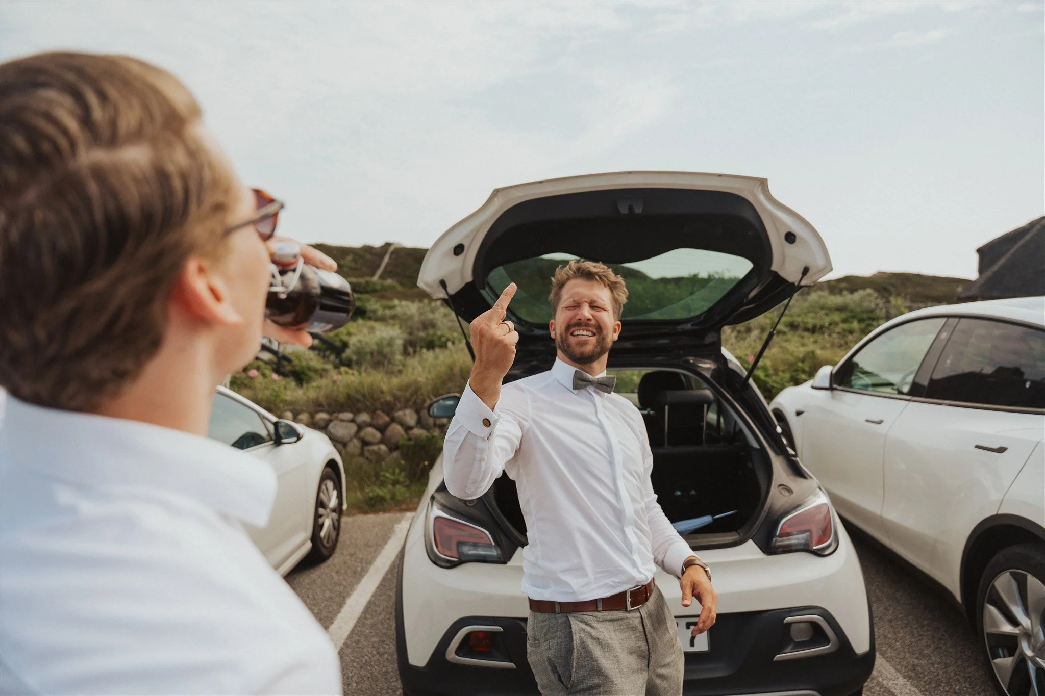 Zwei Männer stehen neben geparkten Autos auf einer Straße, einer steht vor dem Kofferraum eines weißen Autos und zeigt dem anderen einen Mittelfinger, während dieser genüsslich ein Bier trinkt.