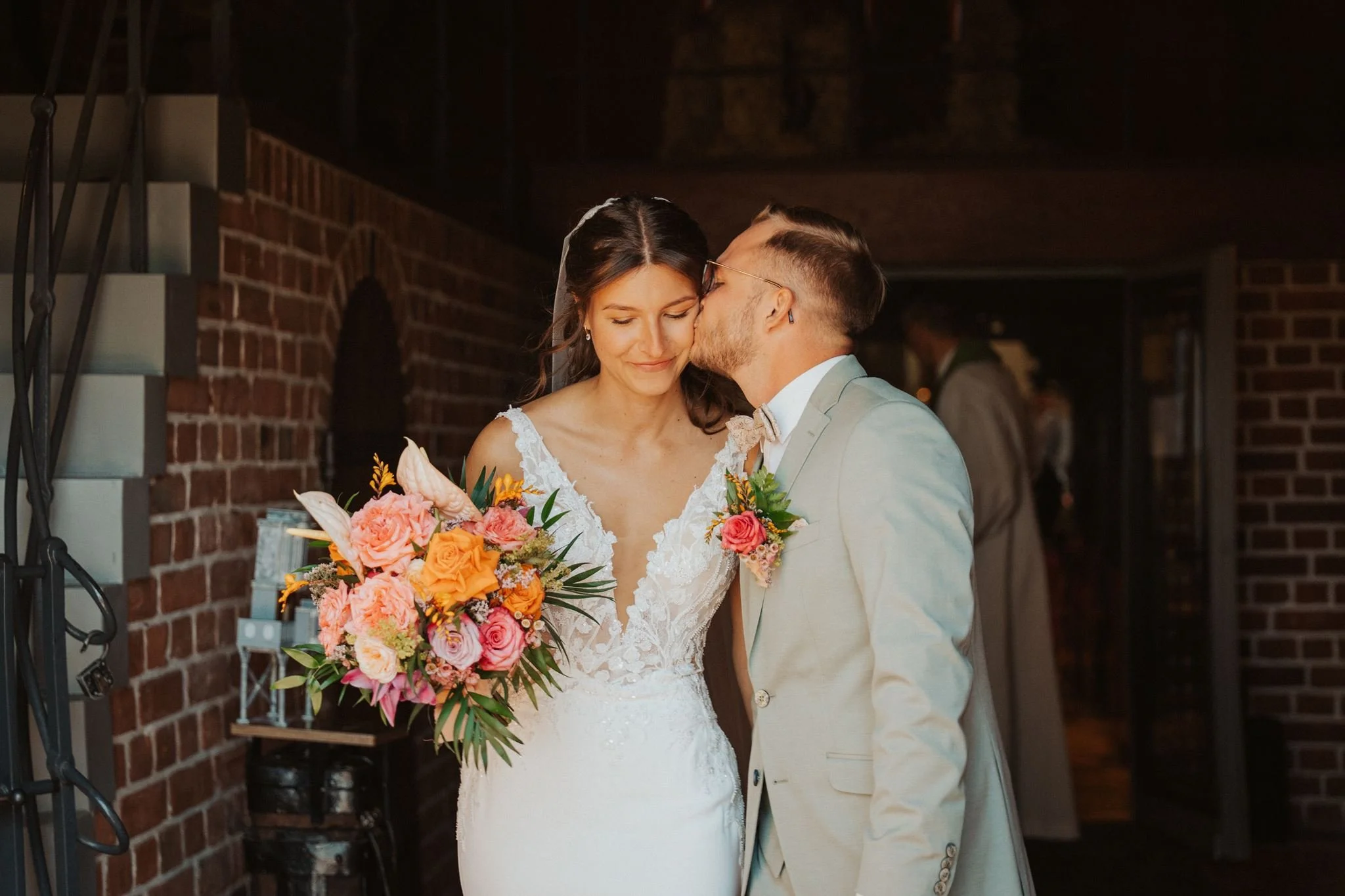 Ein Brautpaar bei einer Hochzeit, der Bräutigam küsst die Braut auf die Wange, beide halten einen großen braunen Blumenstrauß, im Hintergrund eine Wand aus roten Ziegelsteinen.
