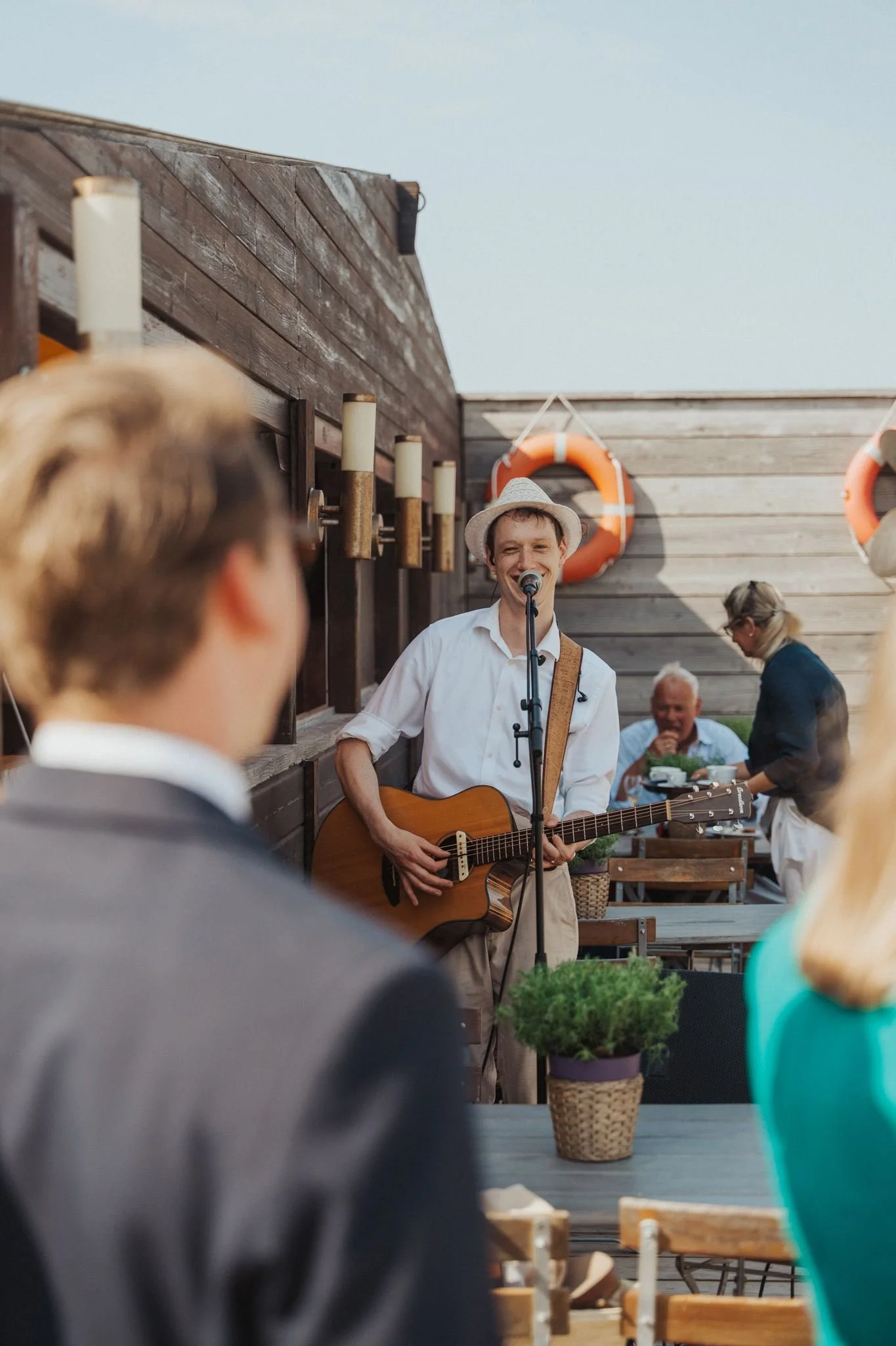 Ein junger Hochzeitssänger spielt Gitarre und singt in einem Außenbereich bei einer kleinen Live-Performance, umgeben von mehreren Menschen, die zuhören und sich unterhalten, mit Holzwänden, Life Buoys und Pflanzen im Hintergrund.