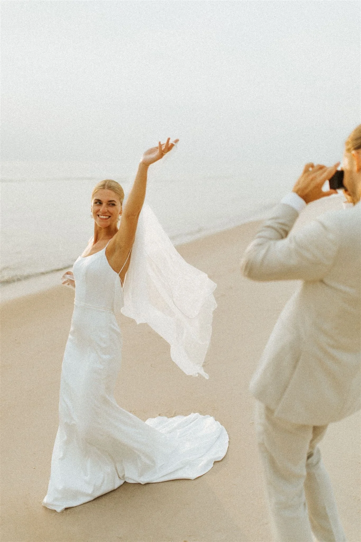 Ein Brautpaar am Strand, die Braut trägt ein weißes Hochzeitskleid und lacht, während der Bräutigam ein Hemd trägt und in die Kamera schaut.
