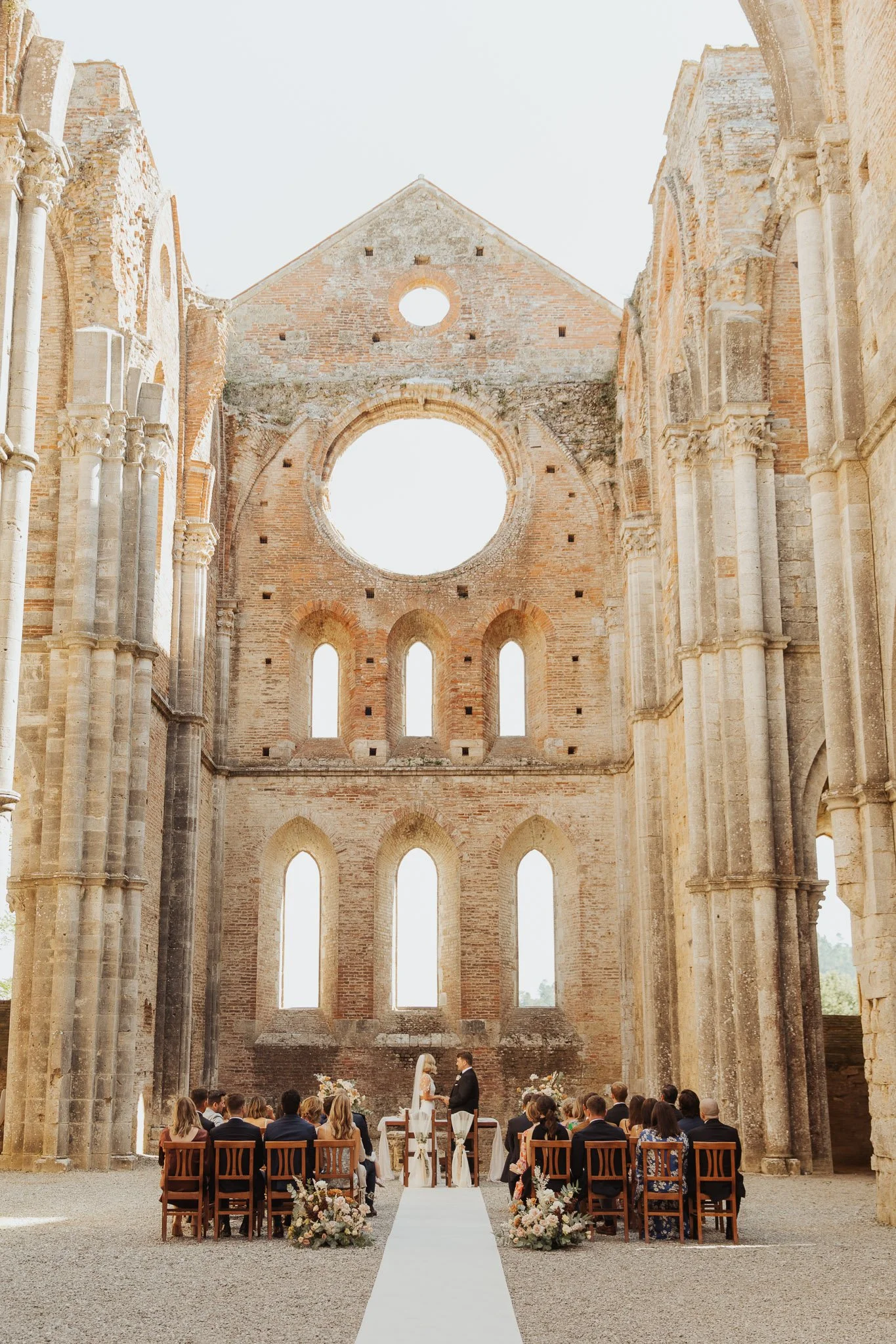 Eine Hochzeitszeremonie findet in den Ruinen einer alten Kirche mit hohen, zerfallenden Ziegelwänden statt. Die Braut und der Bräutigam stehen sich gegenüber, während die Gäste auf Holzstühlen ihnen zusehen.