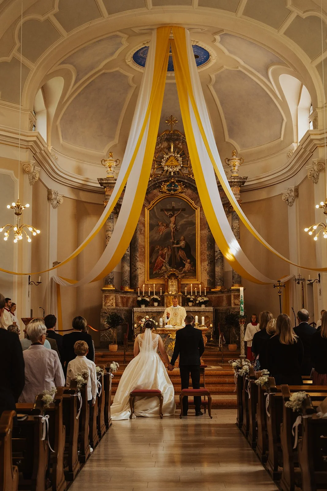 Hochzeit in einer Kirche mit Braut und Bräutigam, die sich die Hand halten, vor dem Altar. Der Priester spricht während der Zeremonie, umgeben von Gästen. Die Kirche ist festlich dekoriert mit gelben und weißen Stoffbahnen und Blumen.
