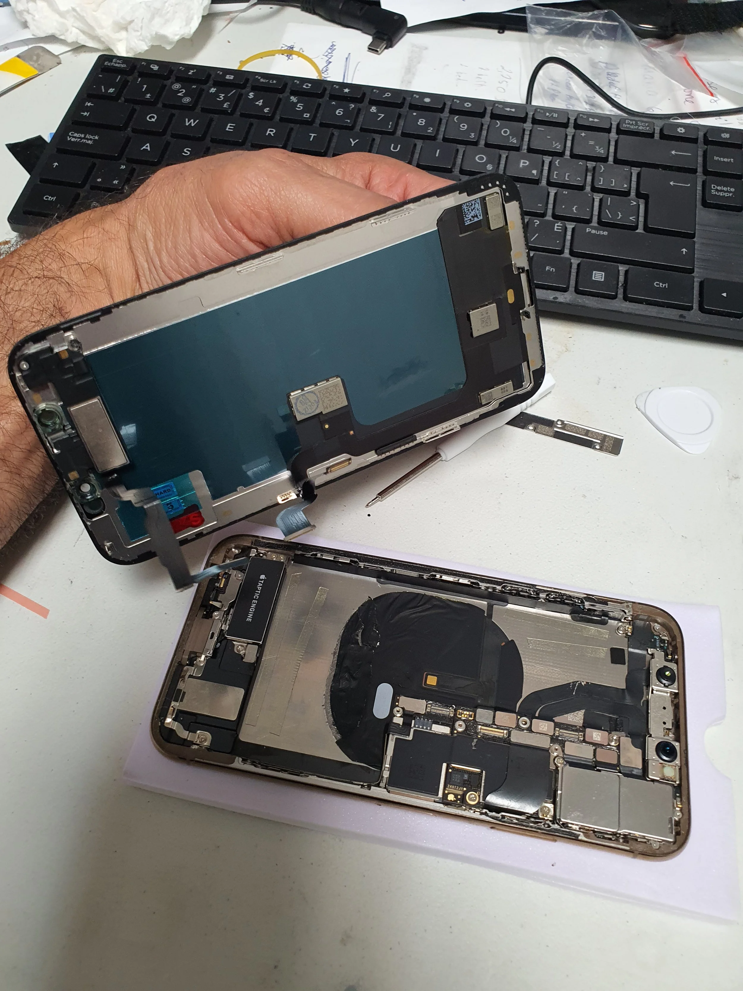Disassembled iPhone showing internal components on a white work surface, with a keyboard in the background.