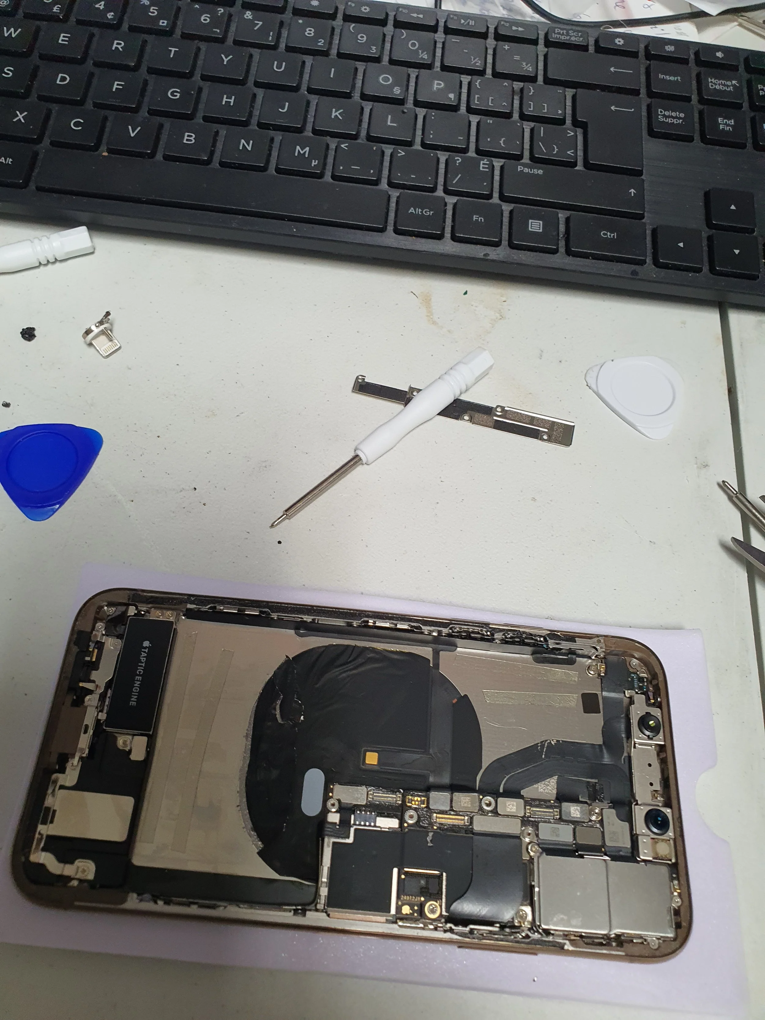 Disassembled smartphone on a white work surface with a keyboard at the top, accompanied by small tools, plastic pry tools, and a SIM card tray.