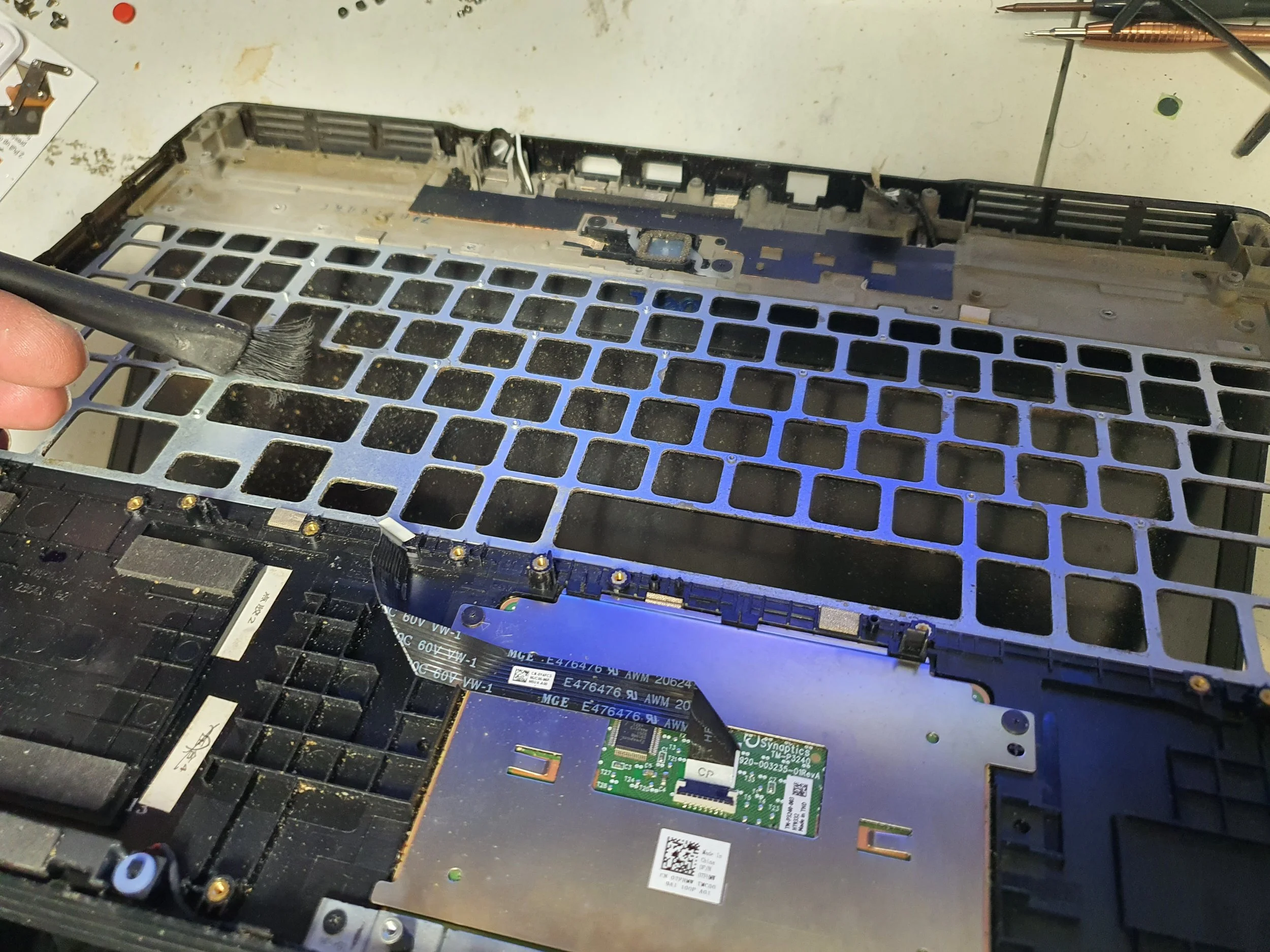 Close-up of a person's hand cleaning the interior of a disassembled laptop with a brush, exposing the motherboard and empty metal frame.
