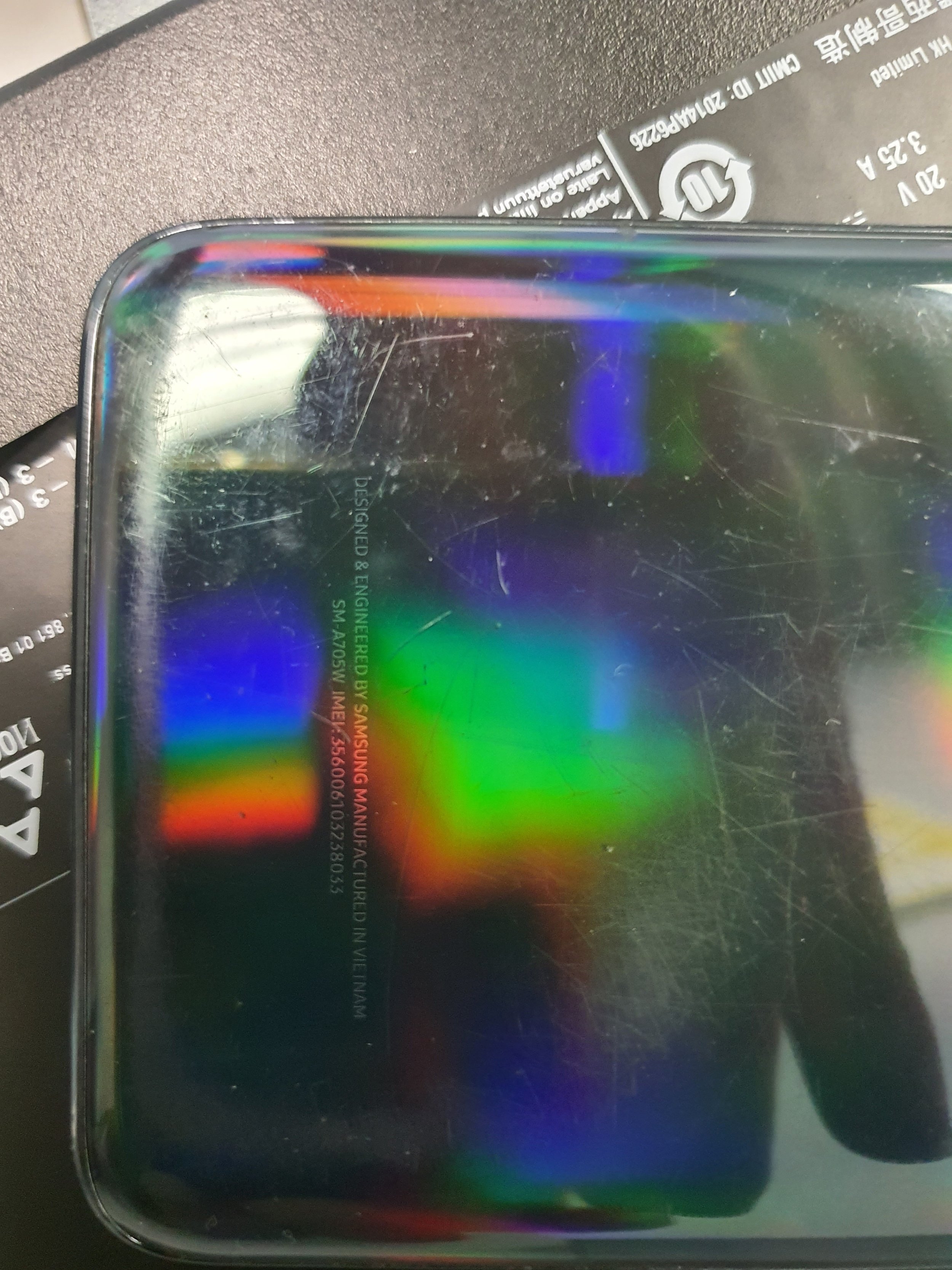 Close-up of a reflective, oily surface of a device, showing scratches and rainbow-colored light reflections. Part of a remote control and a black packet are visible in the background.