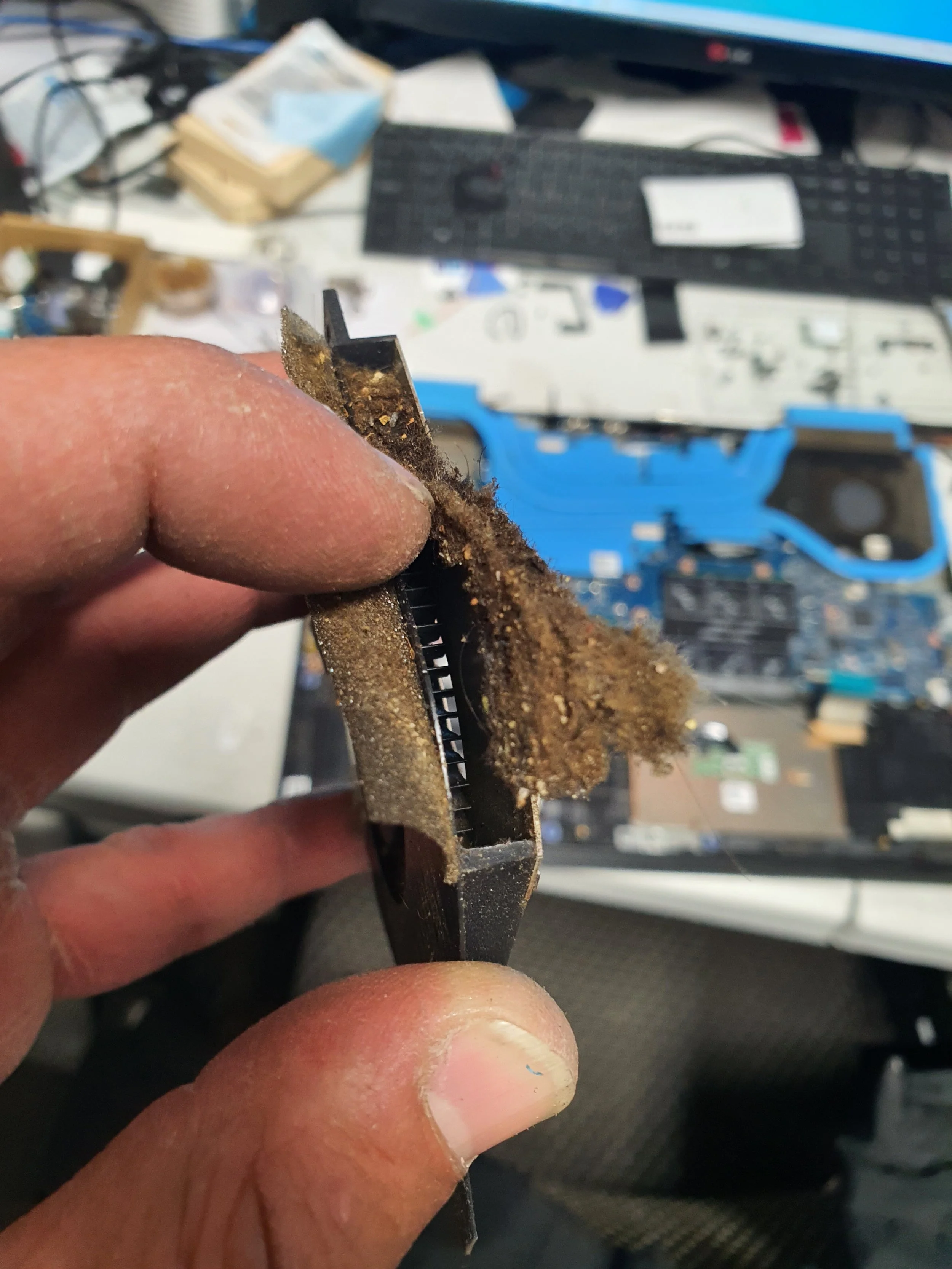 Close-up of a person's hand holding a dirty, dust-covered electronic component, possibly a circuit board or connector, with a cluttered workspace and disassembled electronics in the background.