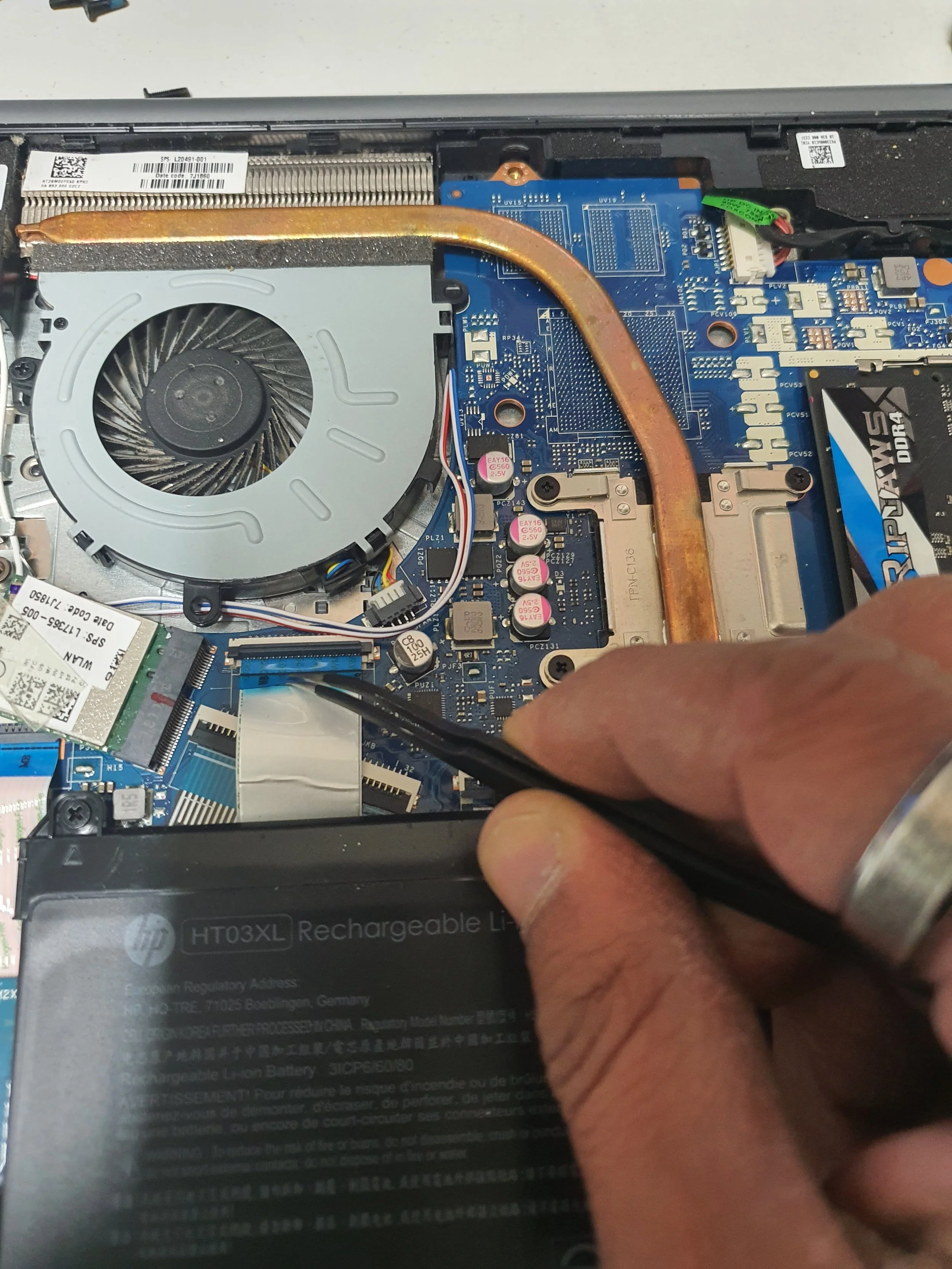 A person using a tool to work on the internal components of a laptop, showing a cooling fan, a copper heat pipe, and a rechargeable lithium-ion battery.