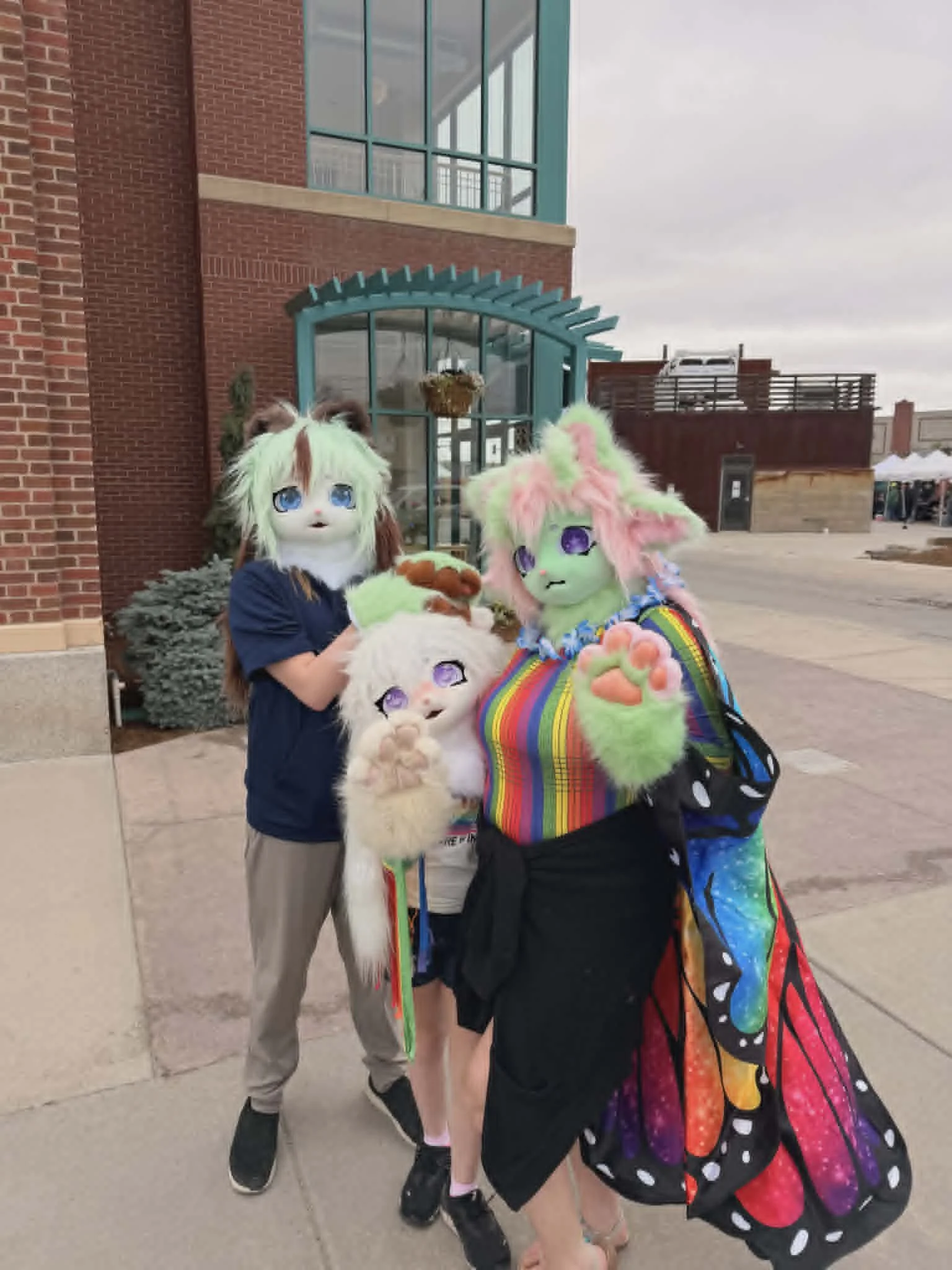 Three people in kemono cat fursuit partials, each with a small pink nose, fluffy heads and paws. One green alien, one white, one mint chip! They're standing just outside the boundary of Casper Pride Fest 2026.