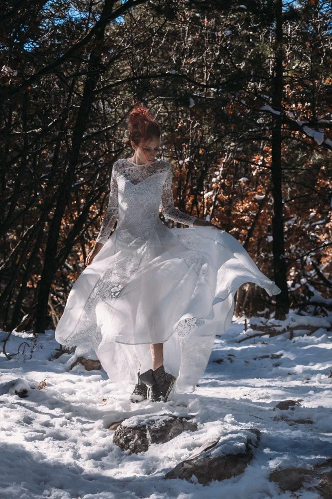 Model Tricia Seaman (Ritter) dancing in the New Mexico snow during an interstate group shoot. Tricia's red hair is up in a messy bun, wearing a white lacey dress and unlaced boots, twirling through snows in front of the trees. @AlexisBerardi