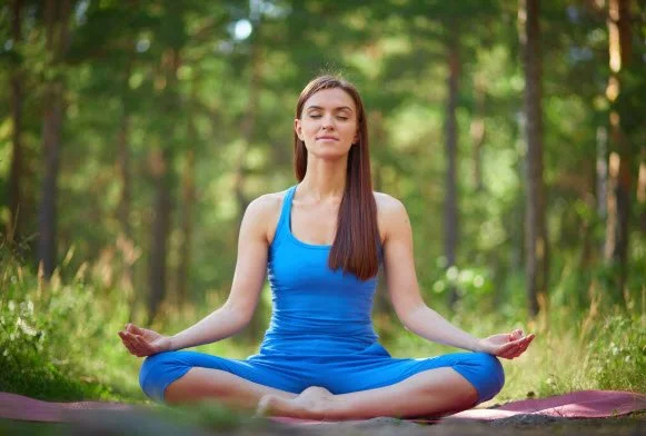 Woman sitting cross-legged in a forest practicing breathing meditation, with her eyes closed and hands resting in a relaxed position.