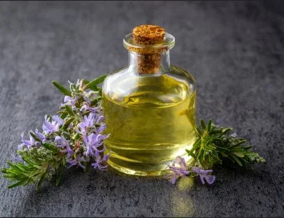 Rosemary oil in a glass bottle surrounded by fresh rosemary sprigs with small purple flowers on a dark surface.