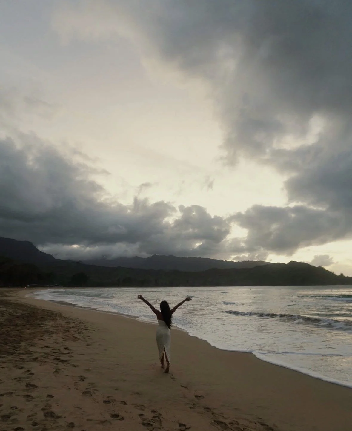 Shaykayla Smith walking on a beach with her arms raised, facing the ocean with cloudy skies overhead and distant mountains in the background.