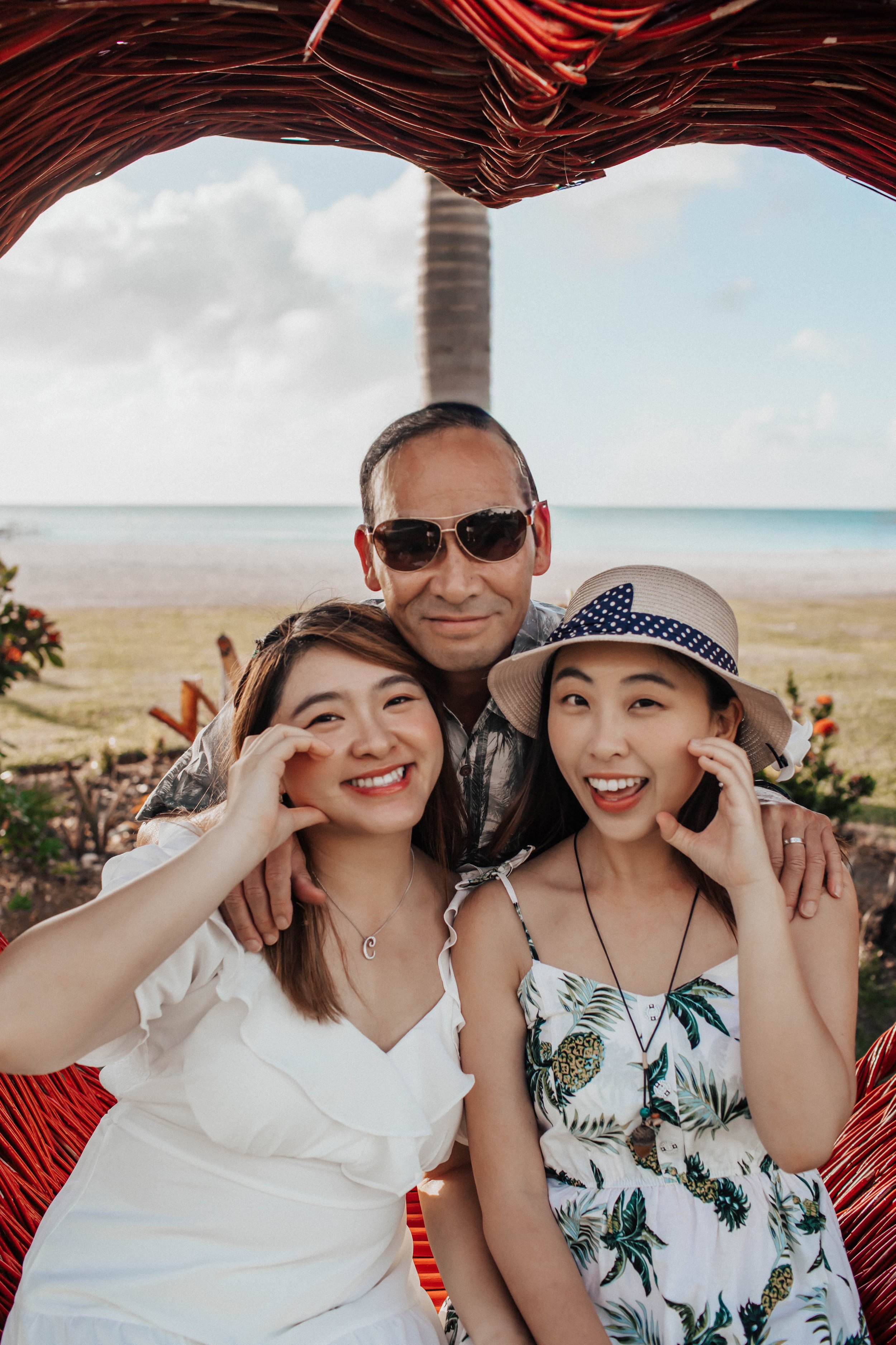 Family of four enjoying a beach outing, smiling and posing for a photo under a woven canopy with the sea and sky in the background.