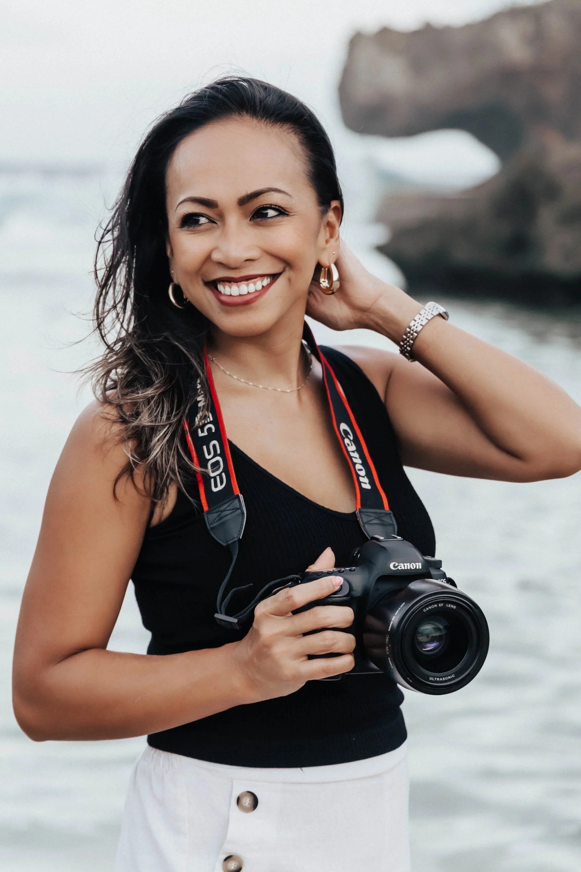 A woman holding a Canon camera around her neck, standing outdoors by the water with rocks in the background, smiling and adjusting her hair.