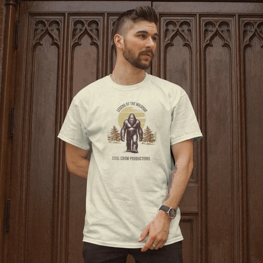 A man with short dark hair, a beard, and a tattoo on his left arm, wearing a white T-shirt with a graphic of a Bigfoot creature and trees, standing in front of a wooden, carved door.