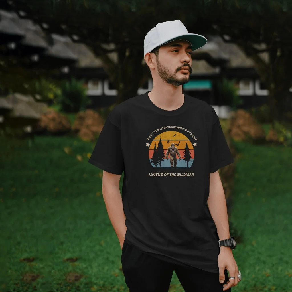 A man wearing a black T-shirt with a graphic of Bigfoot, trees, and a sunset, standing outdoors with a green grass background and dark foliage, looking to the side.