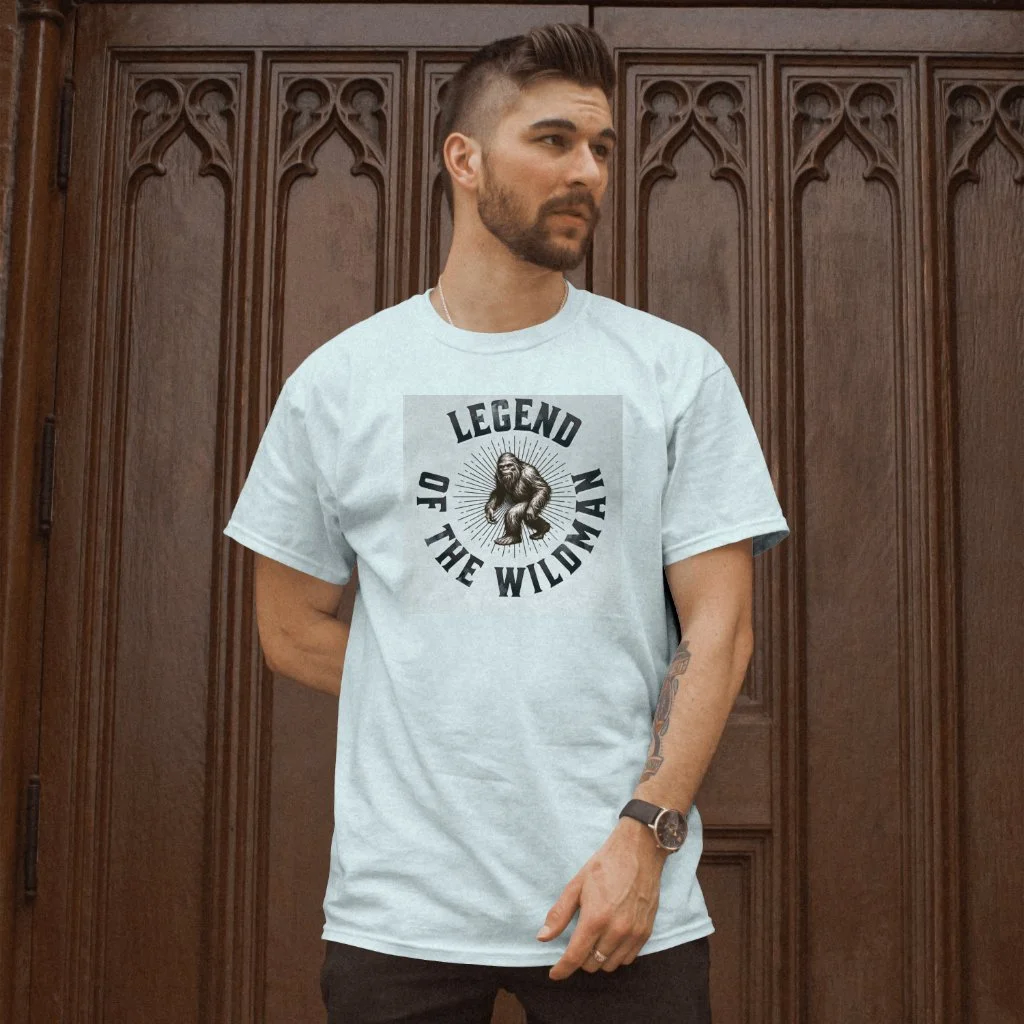 A man with short brown hair, a beard, and a tattoo on his left arm, wearing a light gray T-shirt with a graphic and text that reads 'Legend of the Wildman', standing in front of a carved wooden door.