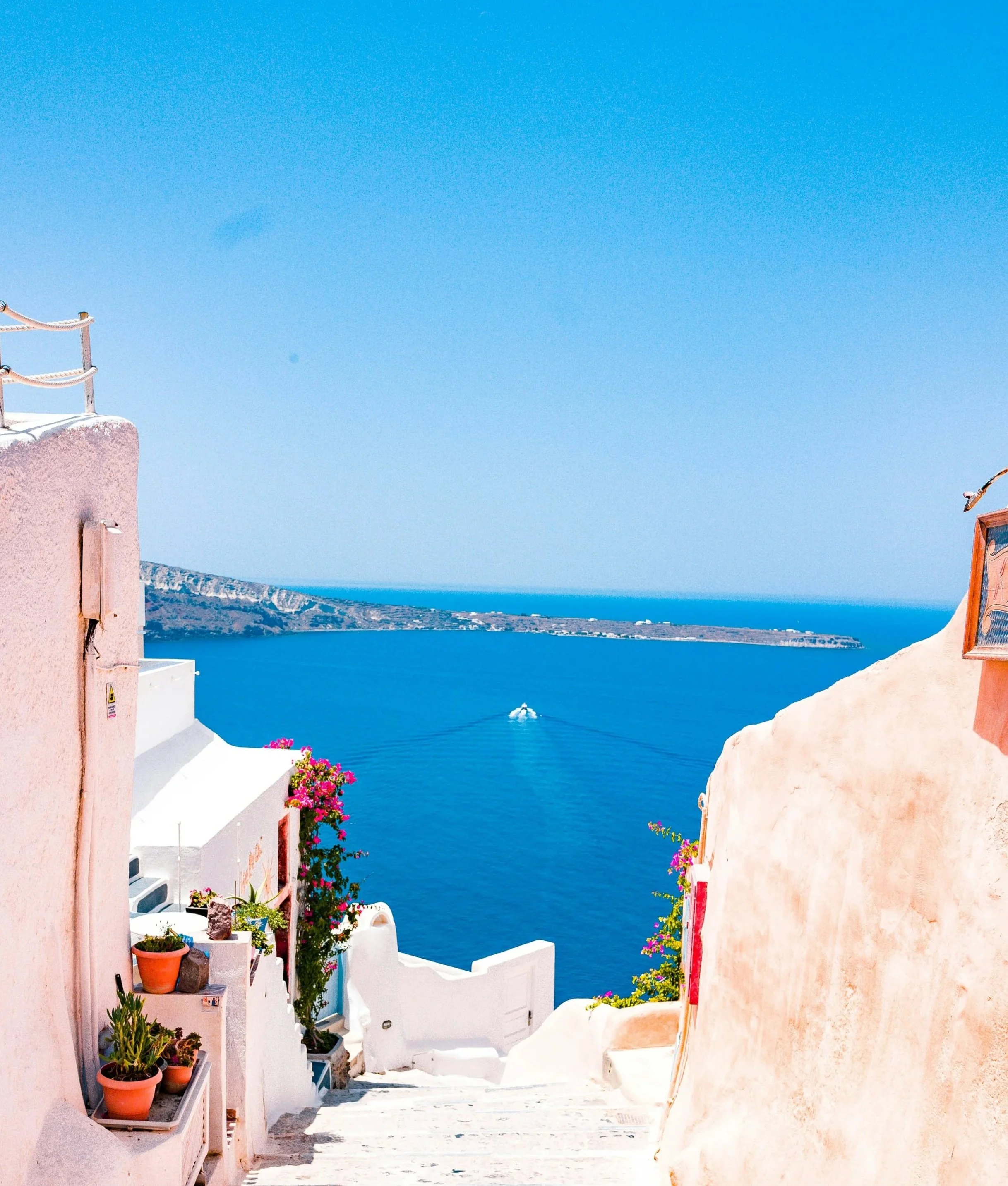 Bright, sunny scene of a coastal village with white and pastel pink buildings, colorful flowers, and a deep blue sea in the background.