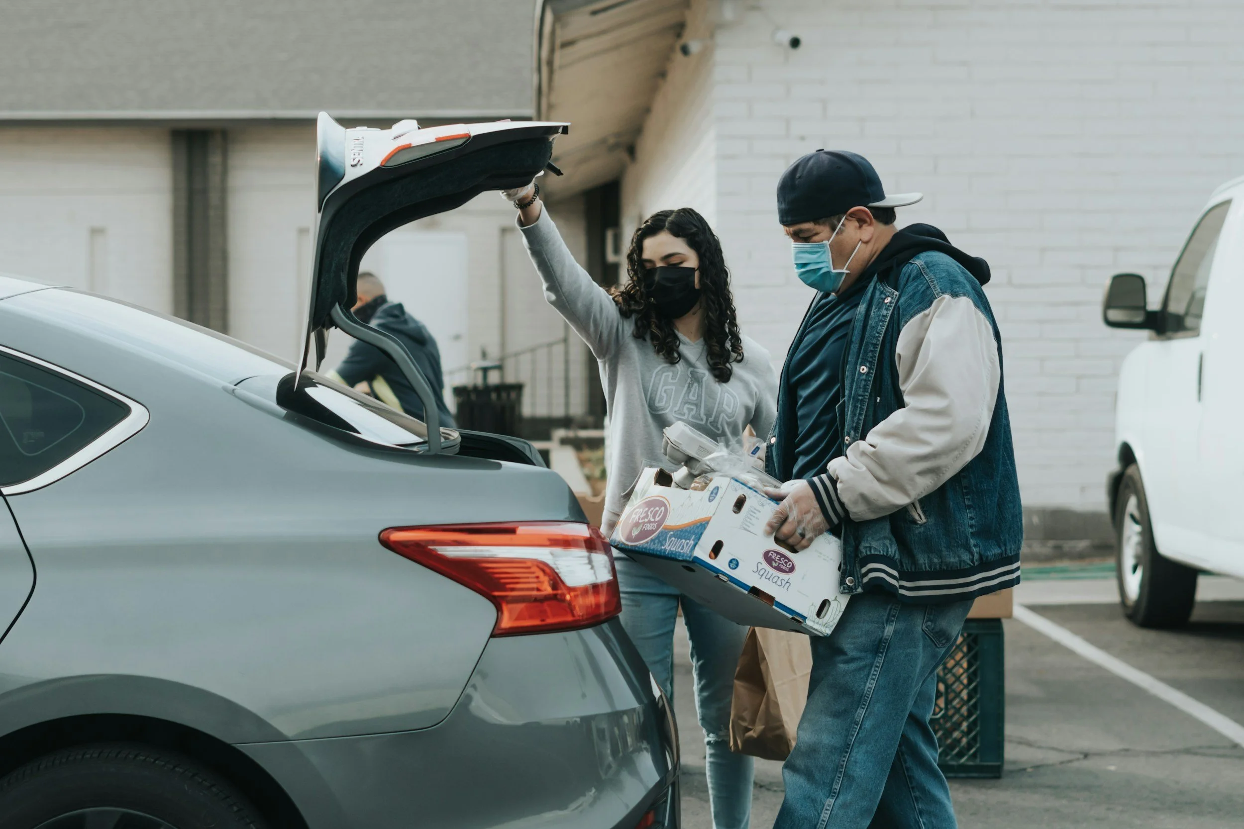 Two people wearing face masks loading groceries into the trunk of a gray car in an outdoor parking lot.