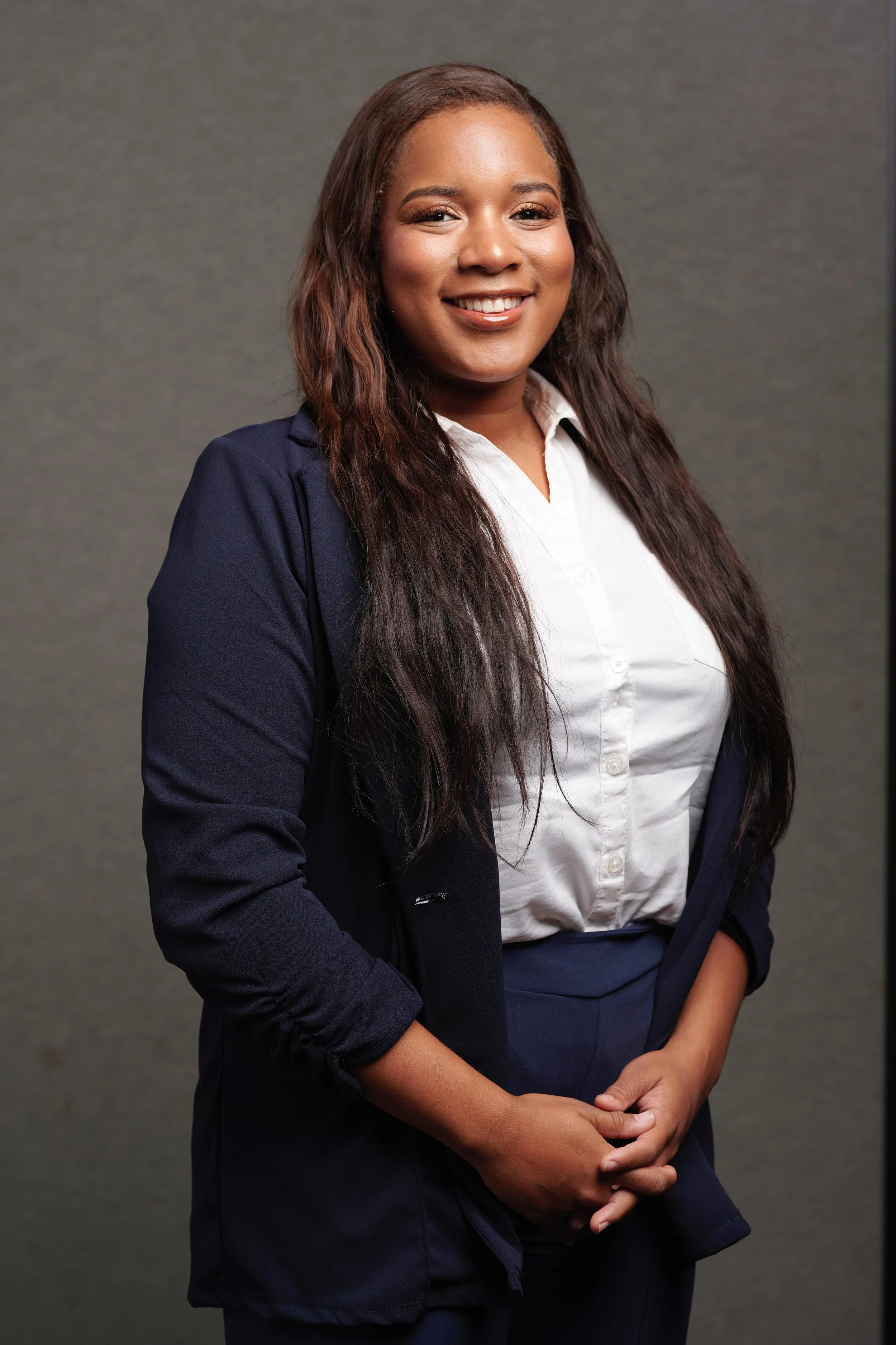 A woman dressed in business attire, smiling, with her hands clasped in front of her, standing against a plain background.