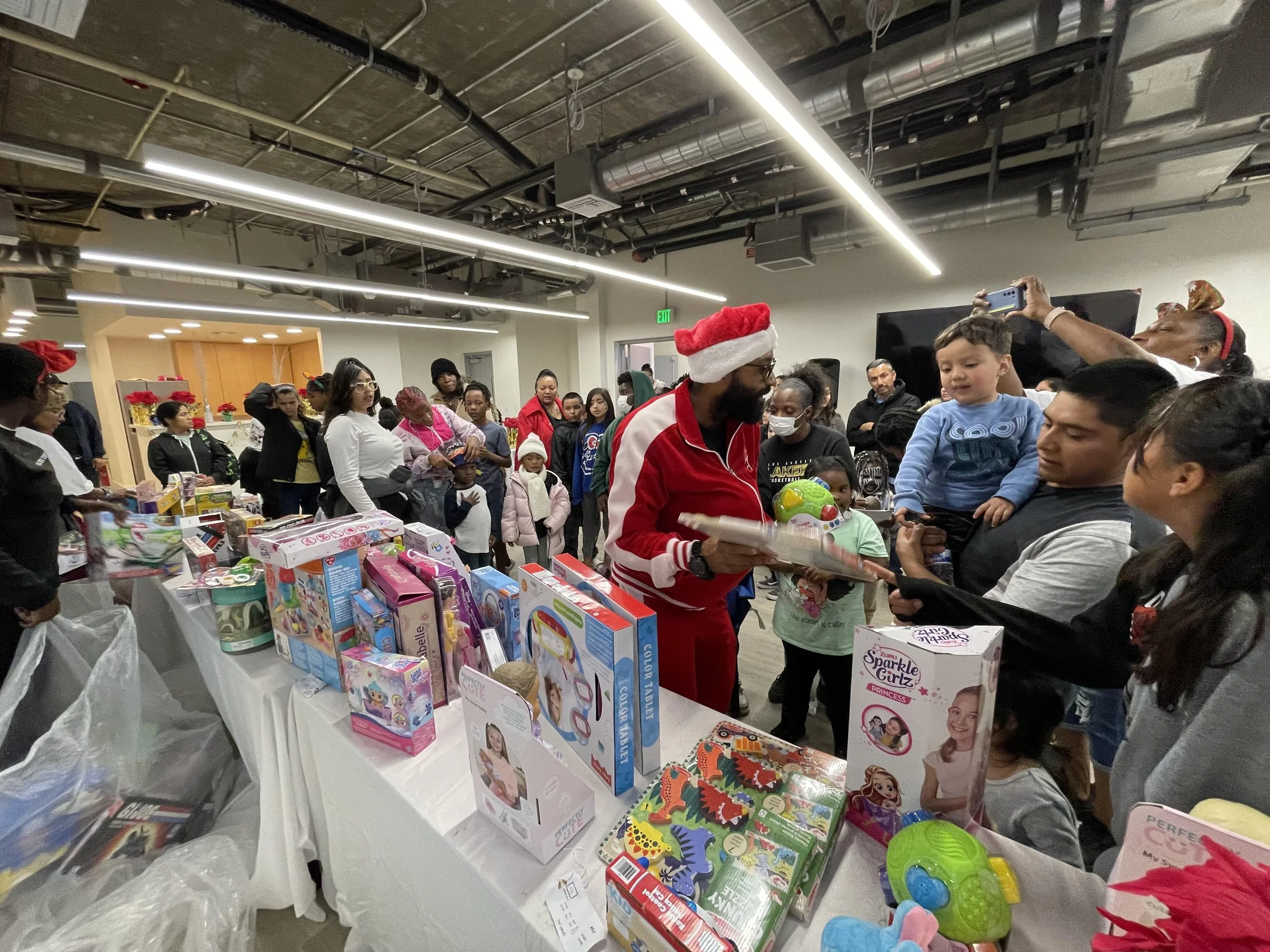 A group of people, including children and adults, gathered indoors at a Christmas-themed event. Many are wearing festive accessories such as Santa hats and headbands. There is a table filled with toys and gifts, and some individuals are holding or selecting presents. The environment has an industrial ceiling with exposed pipes and lighting, and holiday decorations such as poinsettias.
