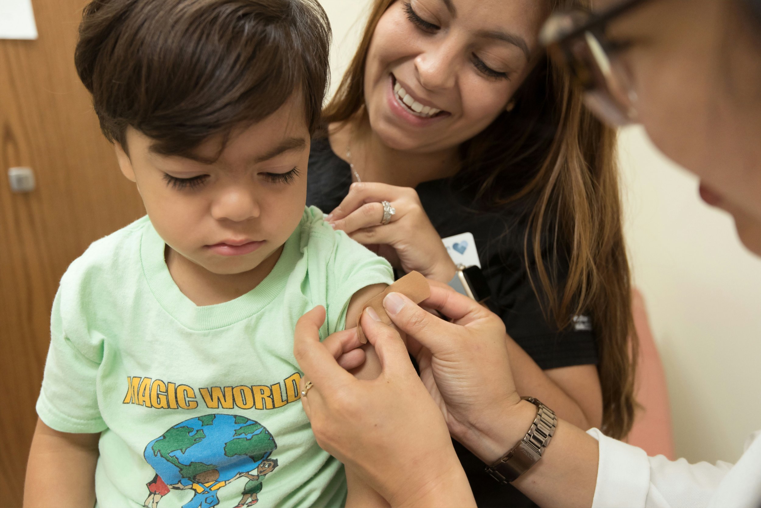 A young boy receiving a vaccination shot on his arm from a healthcare worker, with a smiling woman observing. The boy is wearing a light green t-shirt with a graphic of Earth and the words "Magic World."