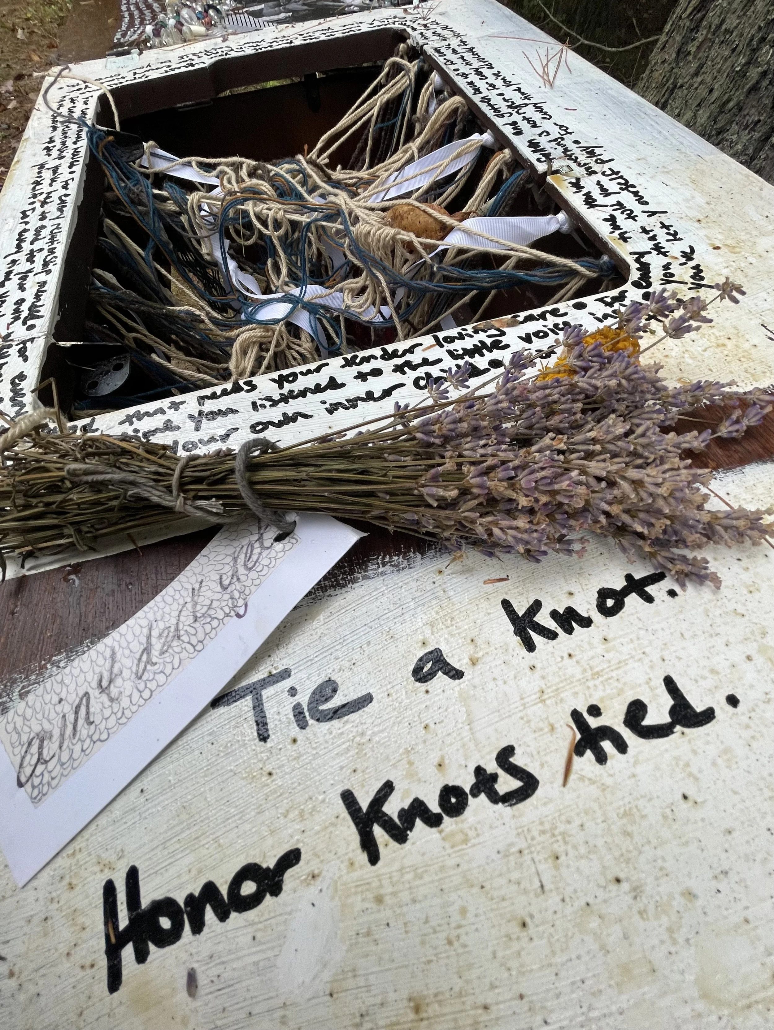 The dirty surface of a white painted waystation features an opening with a tangle of yarn and ribbons. The text "tie a knot. Honor knots tied" is written on the surface in black marker