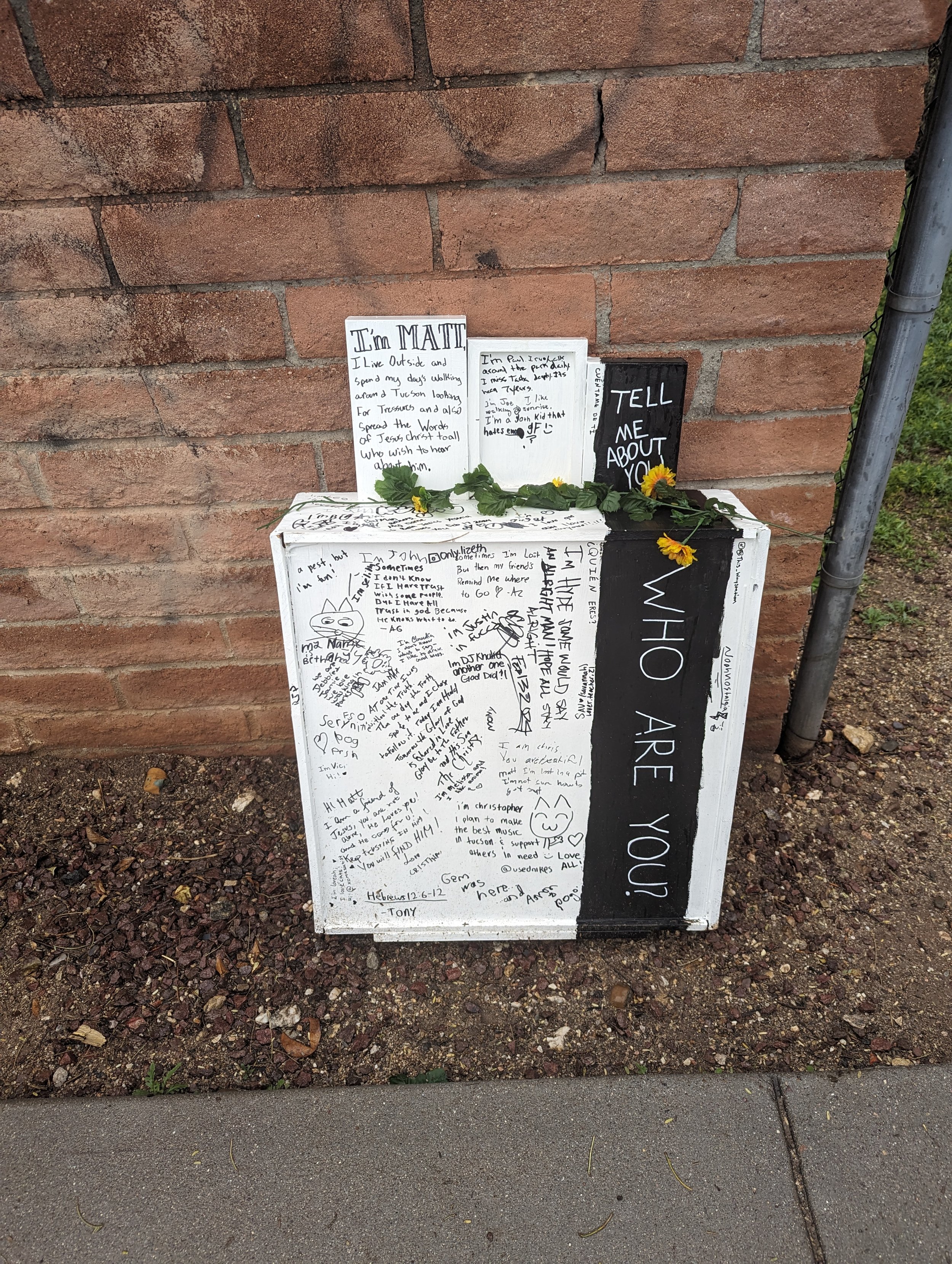 A white waystation covered in writing, on the ground leaning up against a brick wall. A garland of silk sunflowers is laying across the top