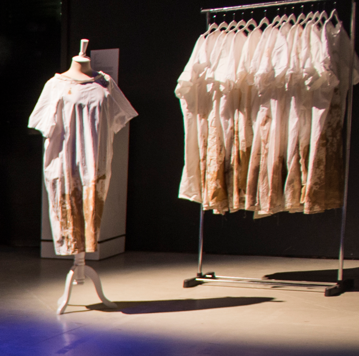 A white hospital gown with brown staining on the bottom half arranged on a dress form with a clothing rack and a number of similar gowns on hangers on the rack.