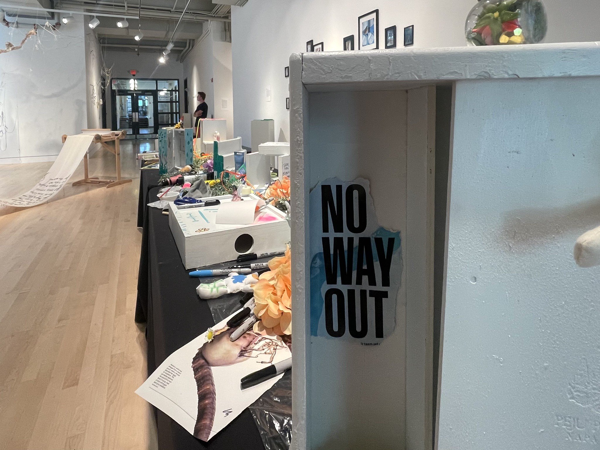 A documentation photo from Amanda Rose's Waystation Generation piece featuring a white waystation with the words "no way out" cut out of a magazine and glued on. A long black table with art supplies is in the background