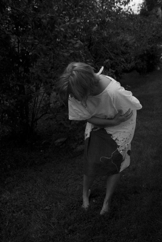 black and white photo of a person wearing a darkly-stained white hospital gown, standing outside barefoot in the grass holding their stomach and doubled over