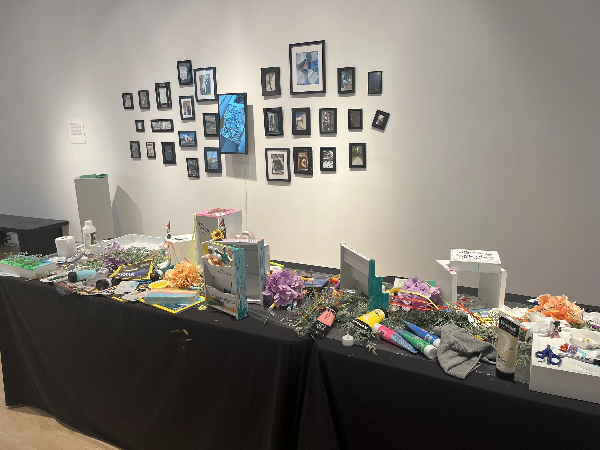 waystation generation piece at the end of the exhibit. There is a long table covered with a black tablecloth and strewn with art and craft supplies, and white-painted pieces with decoration. In the background are framed photos and a digital screen