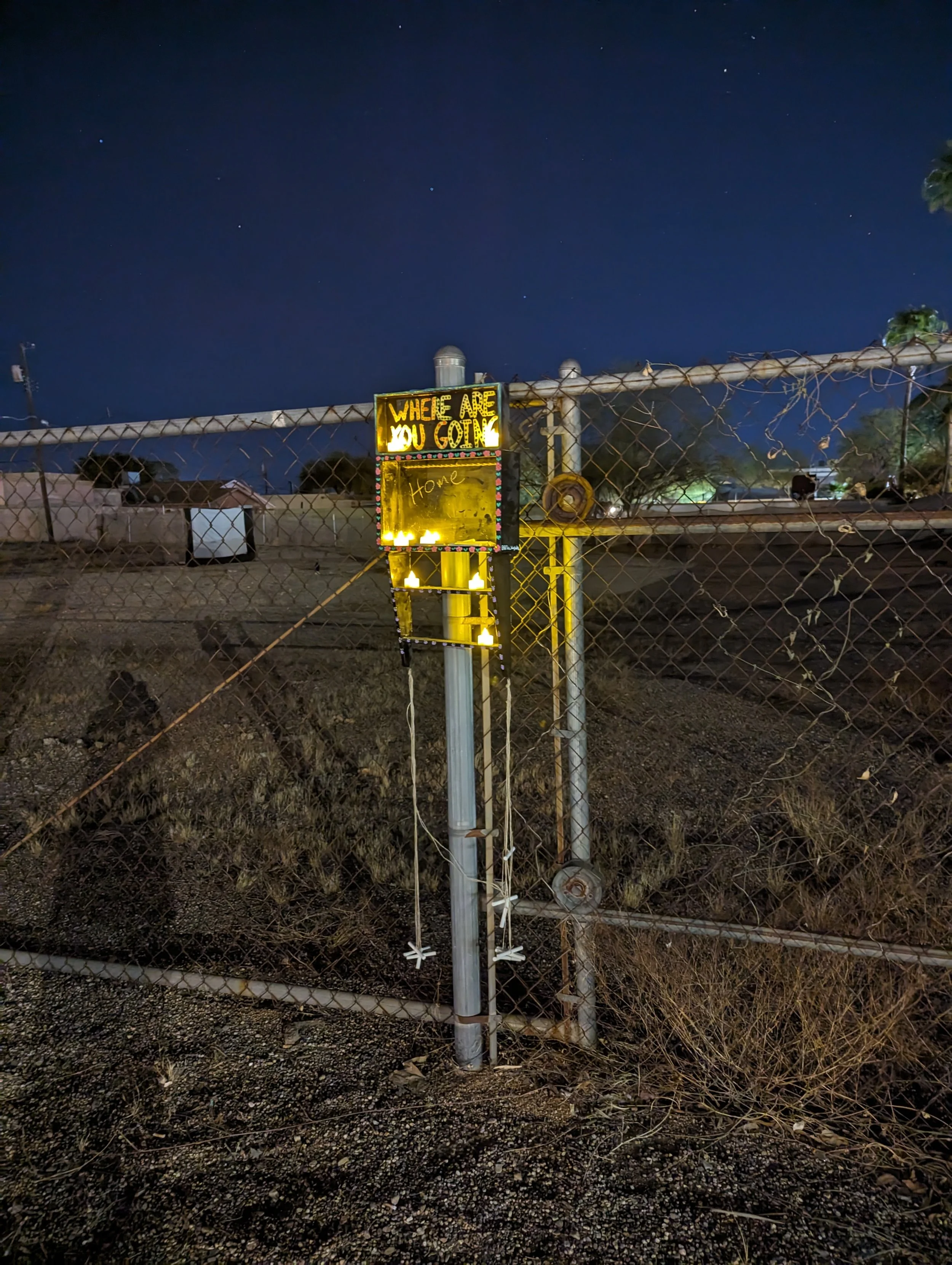 A small black waystation attached to a chain link fence in the middle of a dirt lot at night. The waystation is illuminated with battery operated tea lights and the words "Where are you going" are visible on the waystation