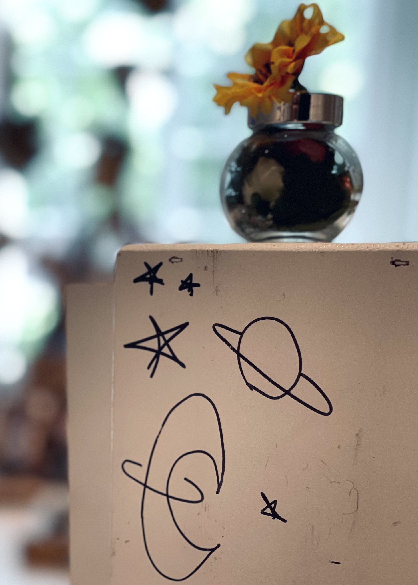 A documentation photo from Amanda Rose's Waystation Generation piece featuring a small glass container with a silk flower resting on top, a white-painted waystation with stars and planets drawn on with black marker