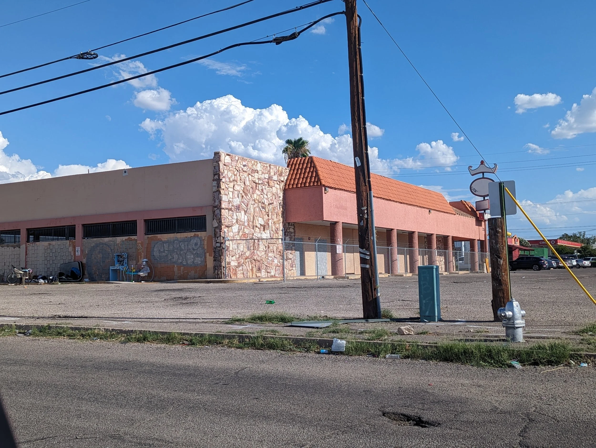 The "Take what you need" waystation placed on the shady side of an abandoned building surrounded by some pieces of baby swings, a bicycle, a plastic lawnchair, and a person sleeping on a bare mattress. It's a sunny day with bright blue sky and clouds
