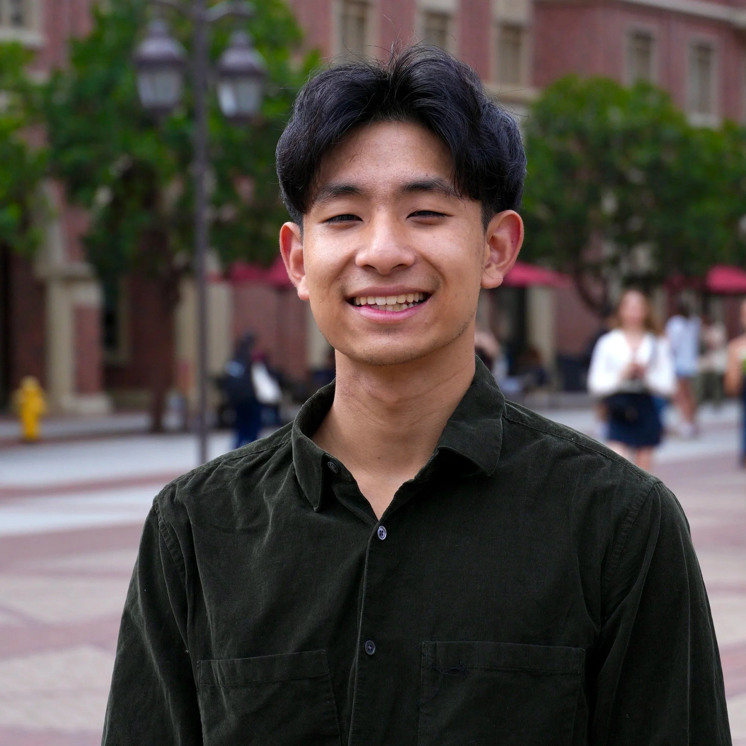 A young man in a blue suit, white shirt, and striped tie, smiling with a brick wall in the background.