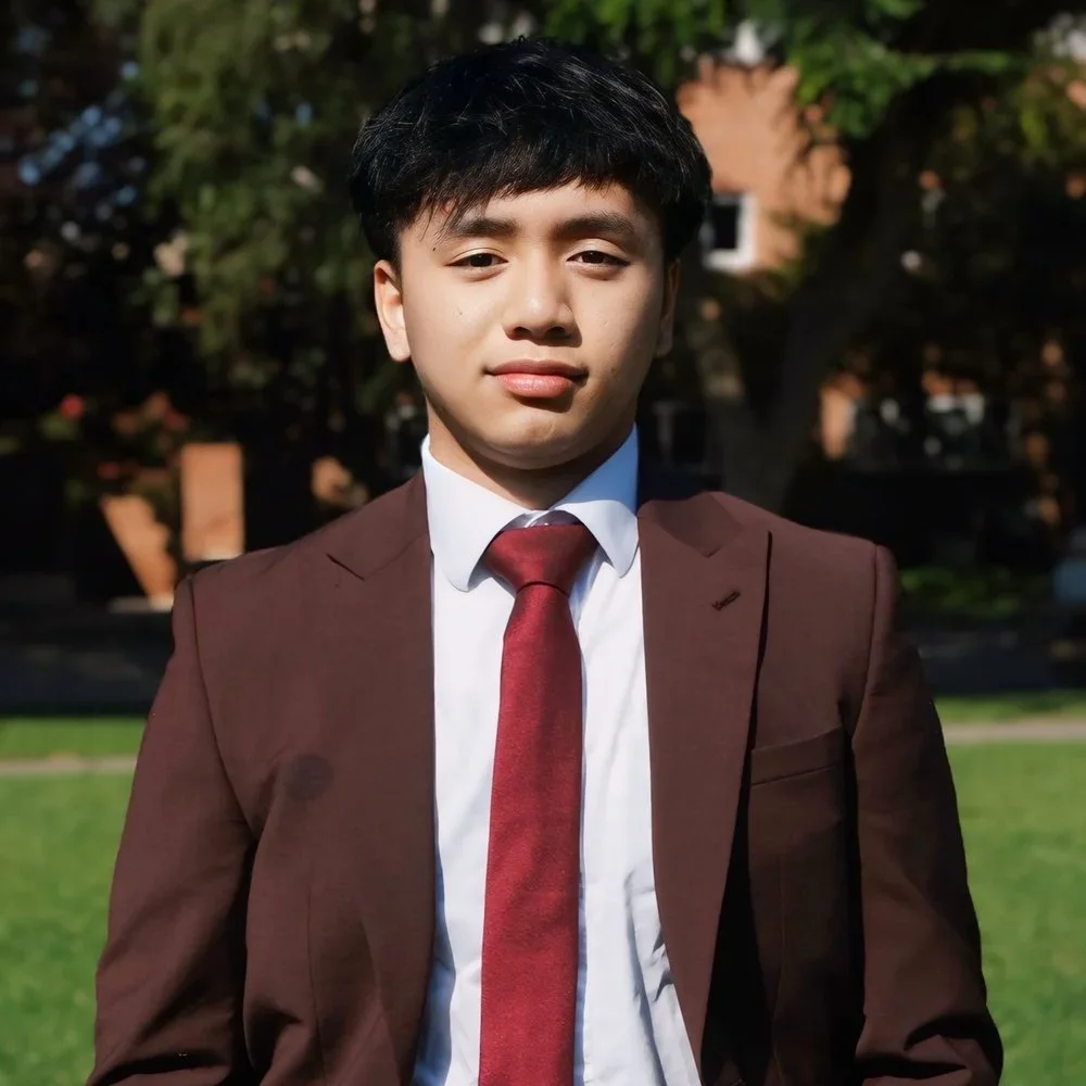 Young man in a gray suit, white shirt, red tie, and black glasses smiling, standing against a beige wall.